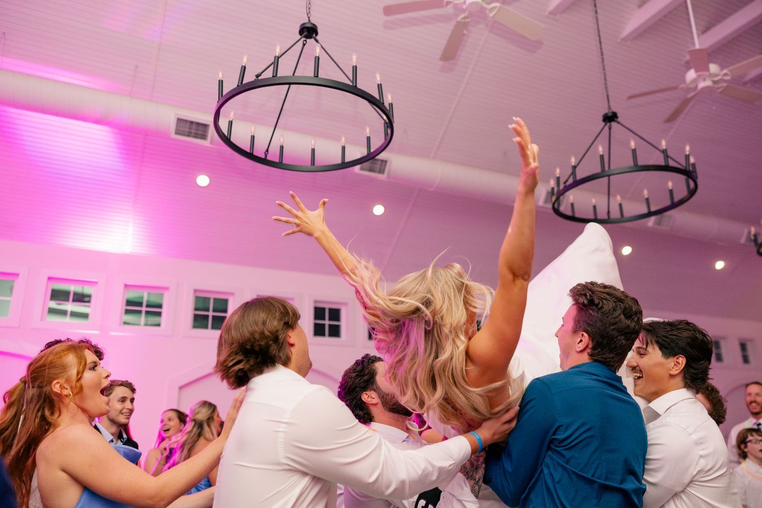 Joyful wedding reception crowd with hands raised dancing under chandelier lights in pink venue.
