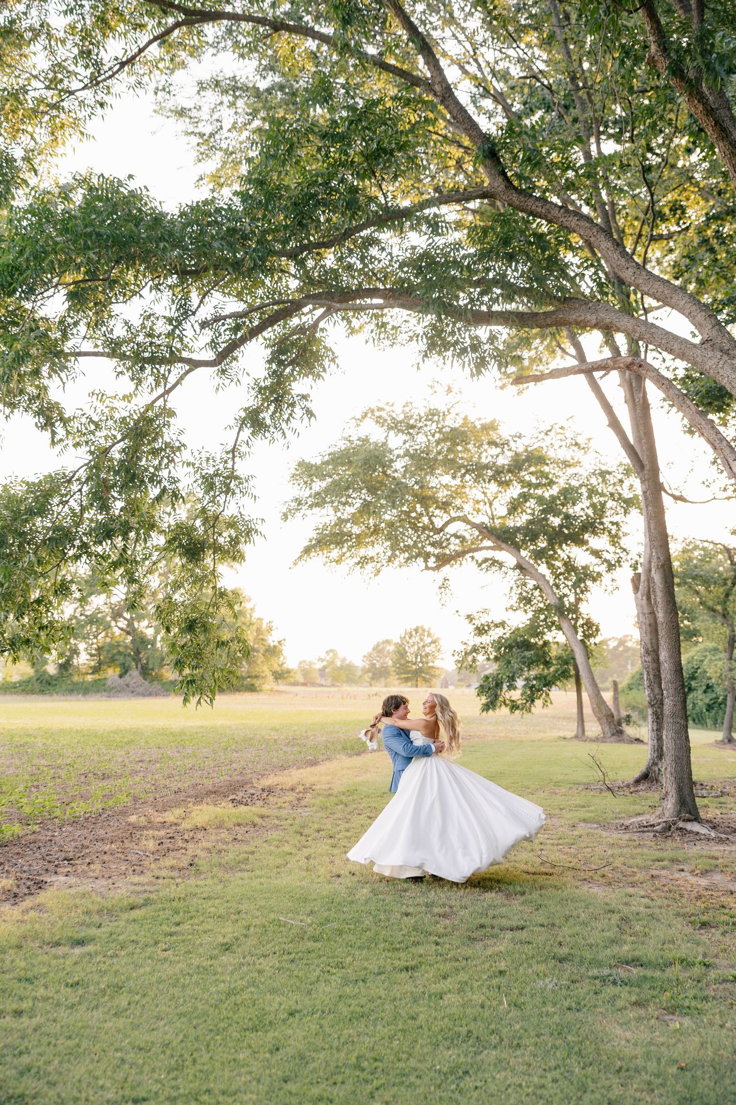 Groom lifts bride in flowing white dress under a large tree at golden hour in a lush green field.