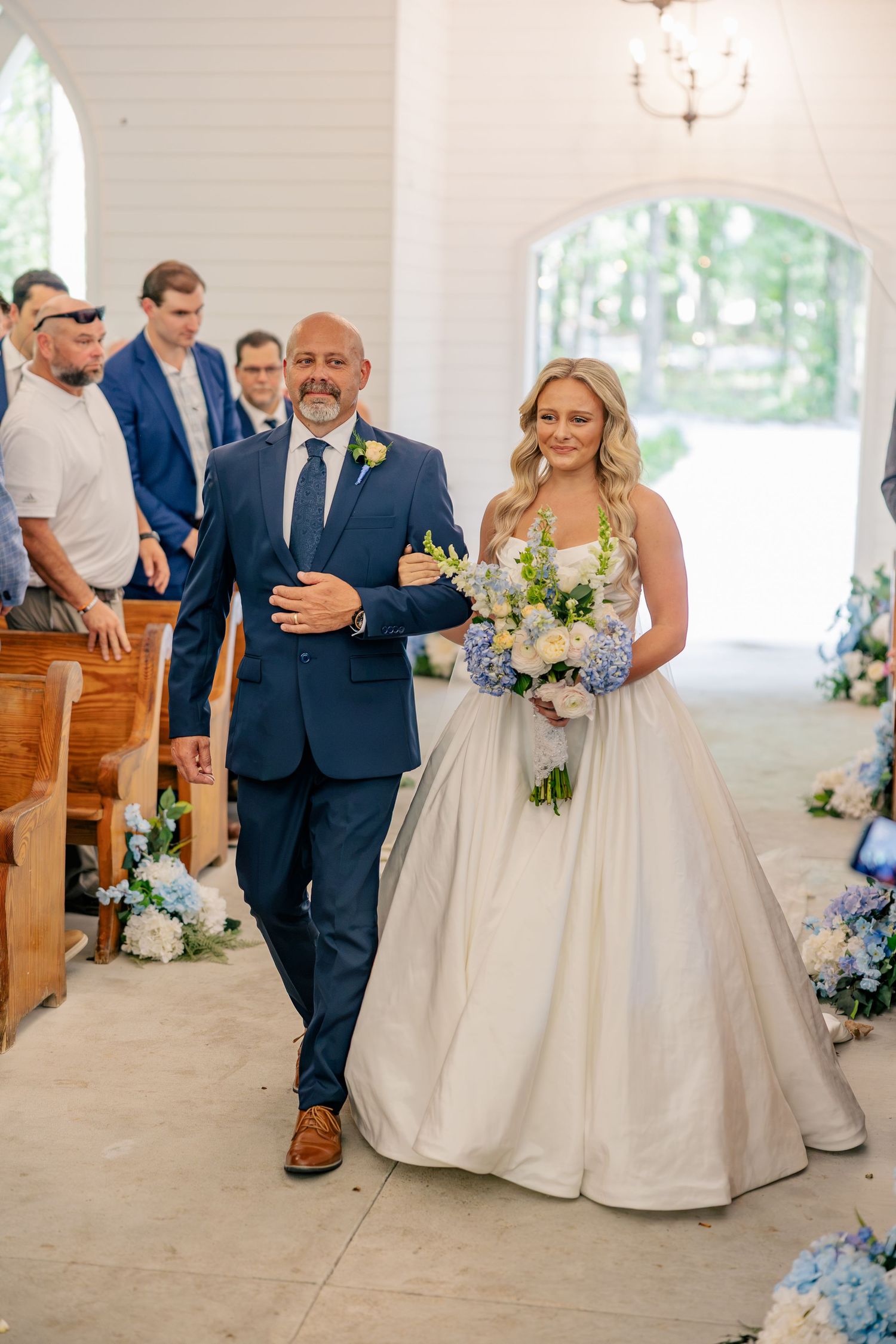 Bride walks down the aisle with escort in navy suit, holding blue and white bouquet in elegant white chapel.