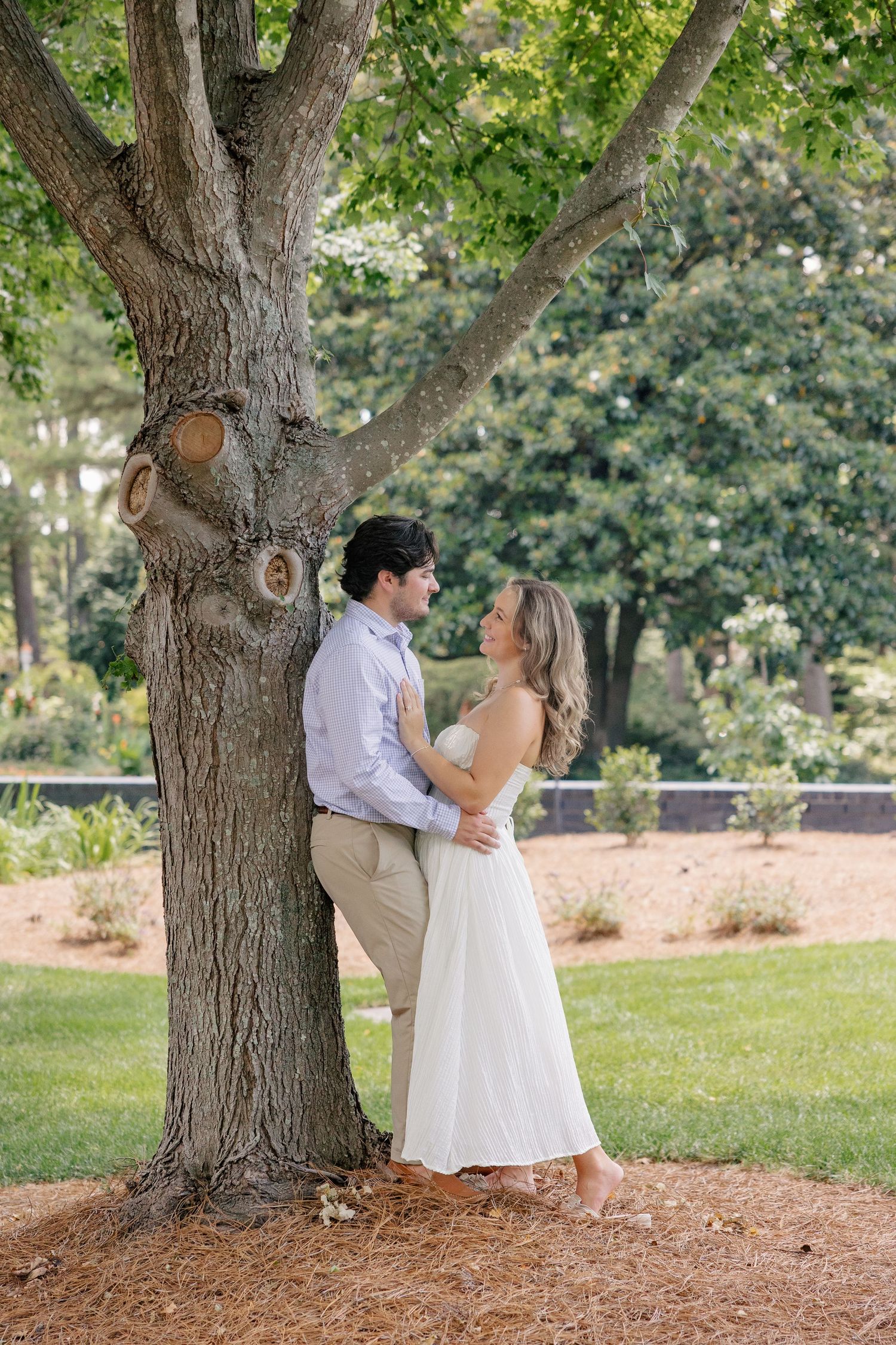 Couple embracing under a large tree in a lush green park, woman in white dress, man in light blue shirt.