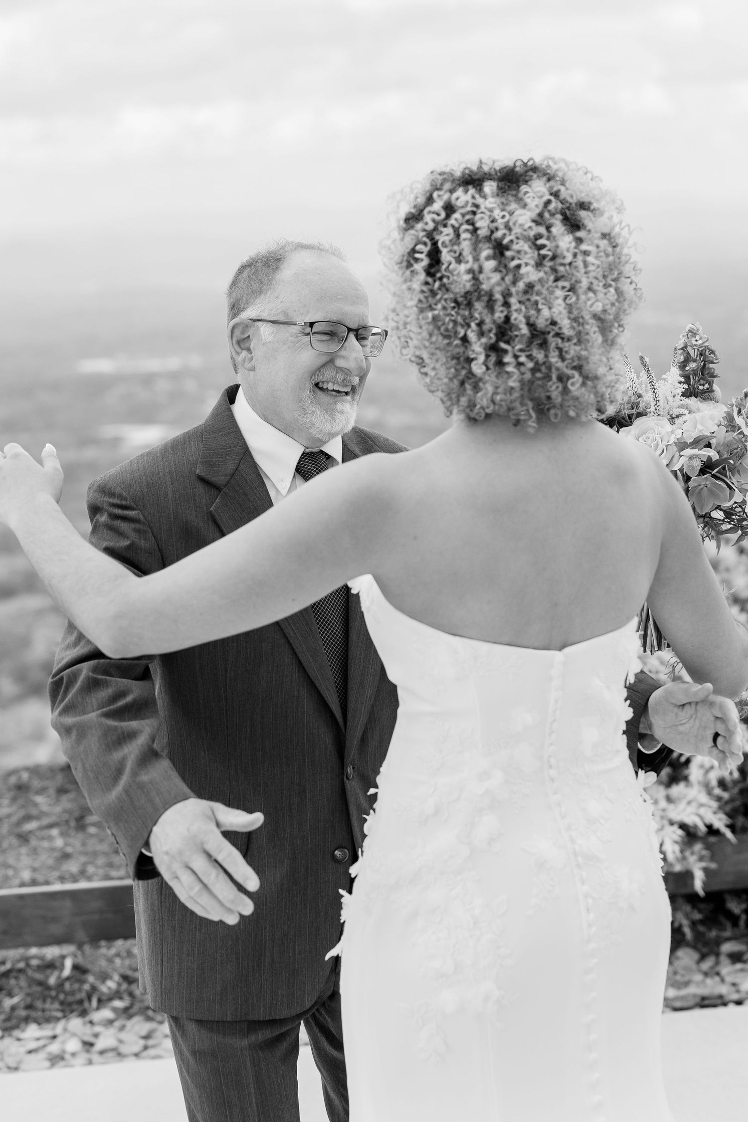 Black and white photo of a bride in a lace gown sharing a joyful first look moment with a smiling man in a suit.