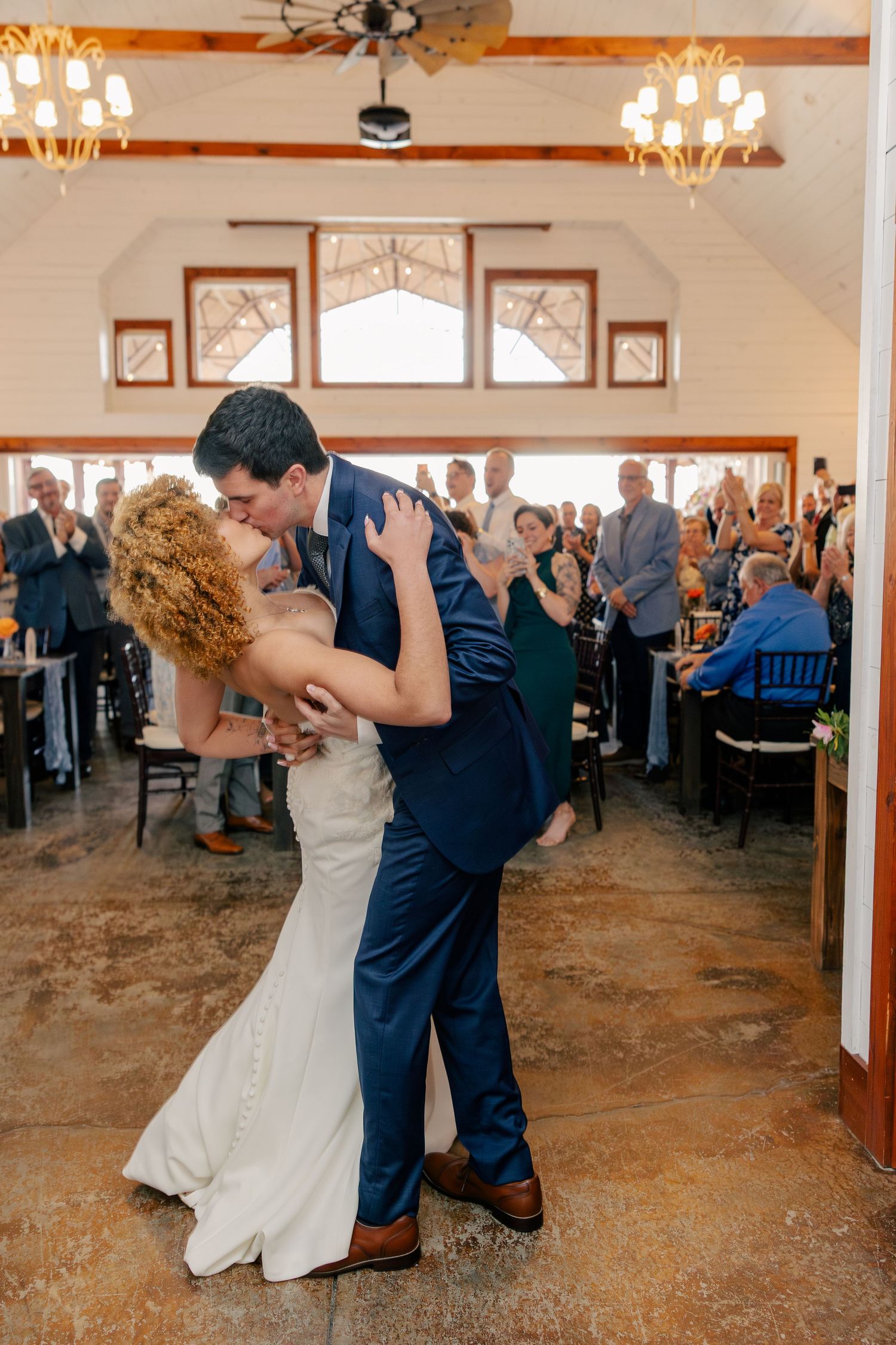 Bride and groom share romantic dip kiss on dance floor surrounded by applauding wedding guests indoors.