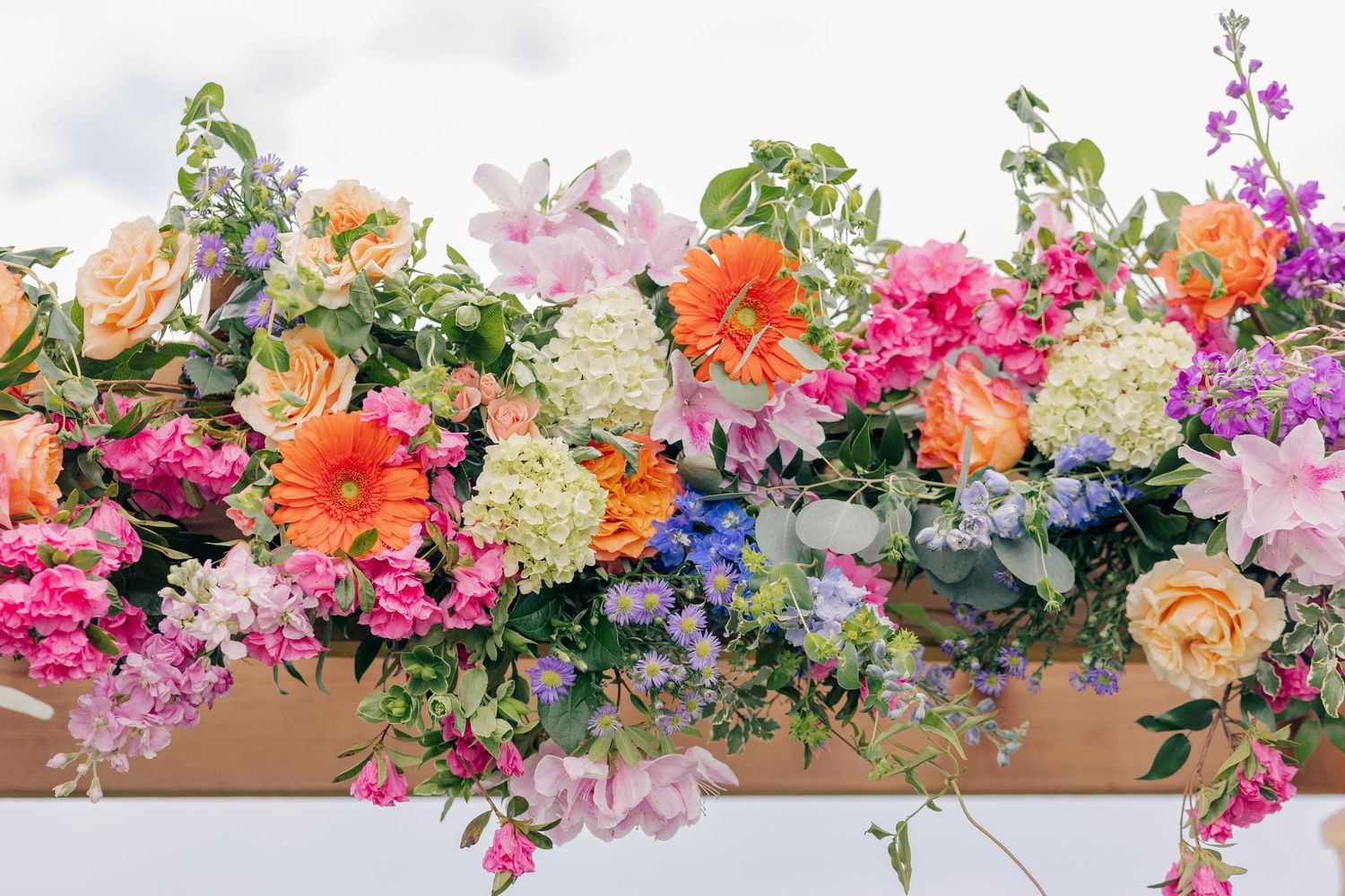 Vibrant wedding arch floral arrangement featuring orange gerberas, pink roses, hydrangeas, and purple blooms.