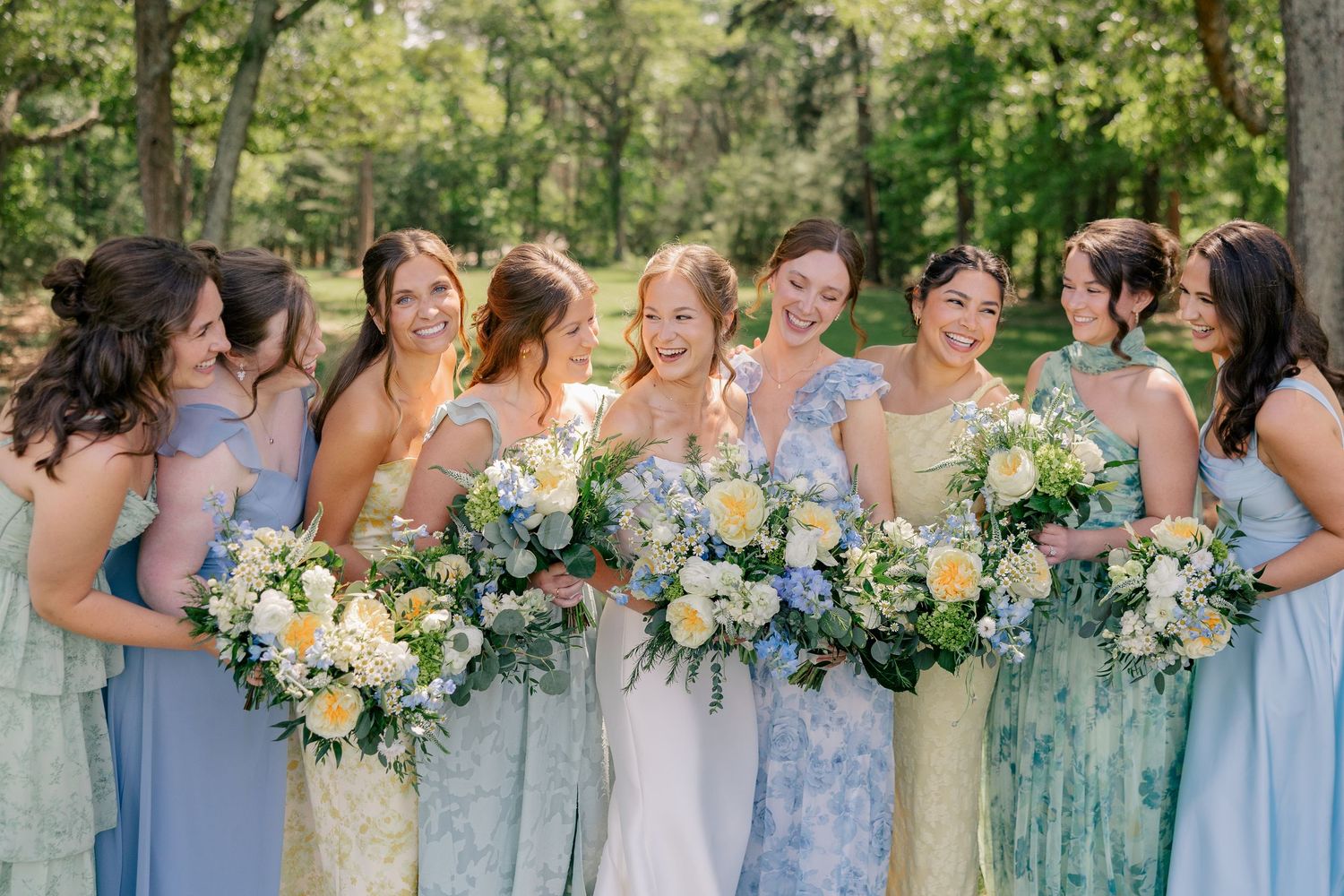 Bridesmaids in mismatched blue and green dresses laugh together while holding white and yellow floral bouquets outdoors.