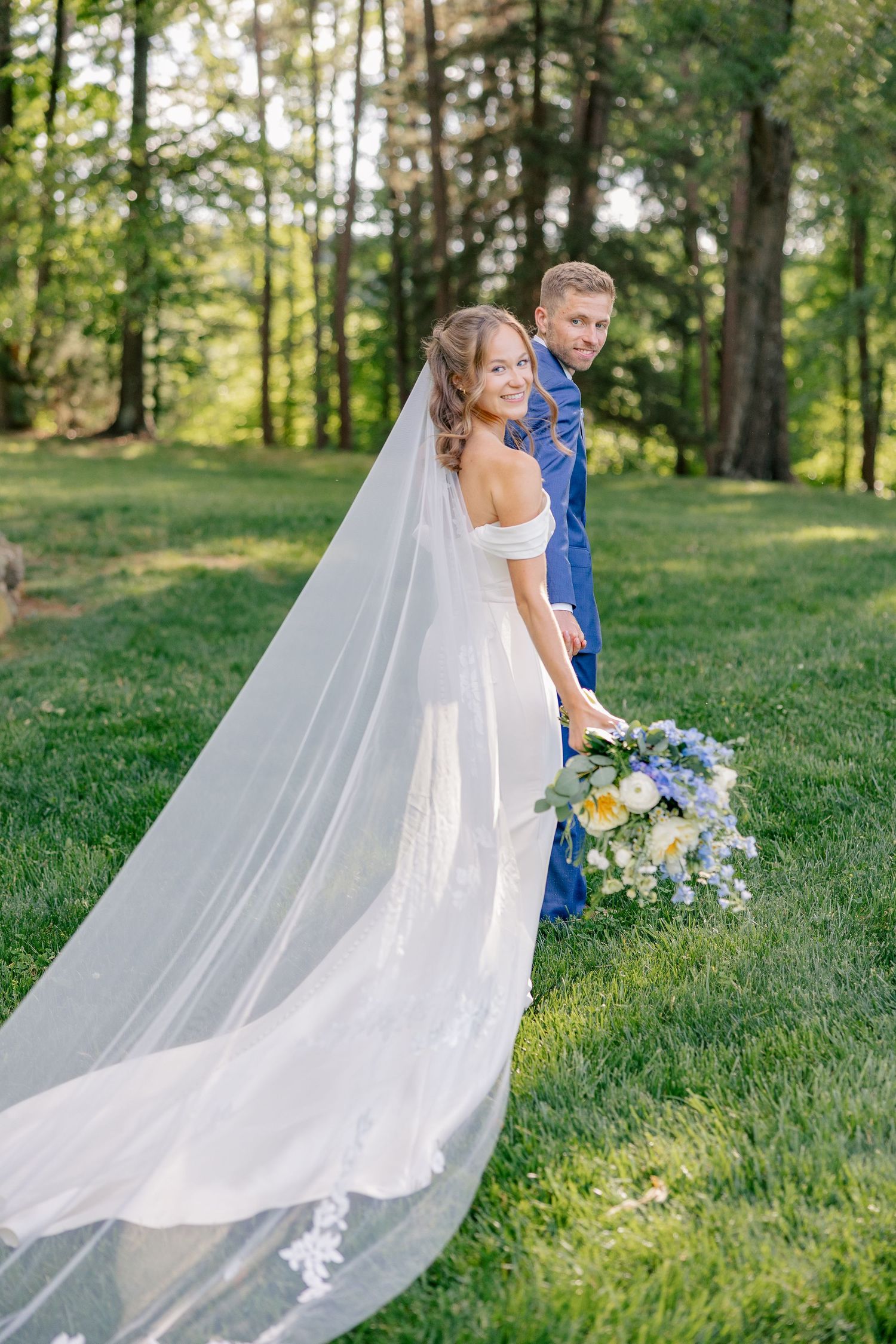 Bride and groom pose together outdoors, her cathedral veil trailing on grass, holding blue and white bouquet.