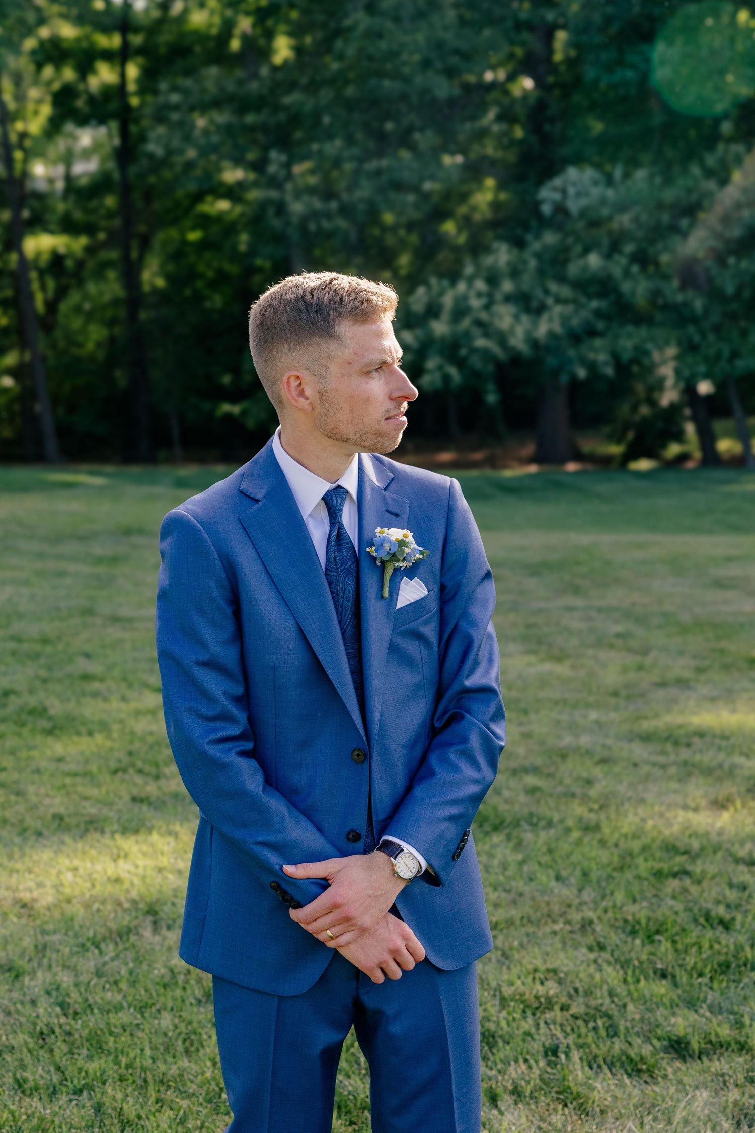 Handsome groom in blue suit with boutonniere stands on grass looking away at outdoor wedding venue.