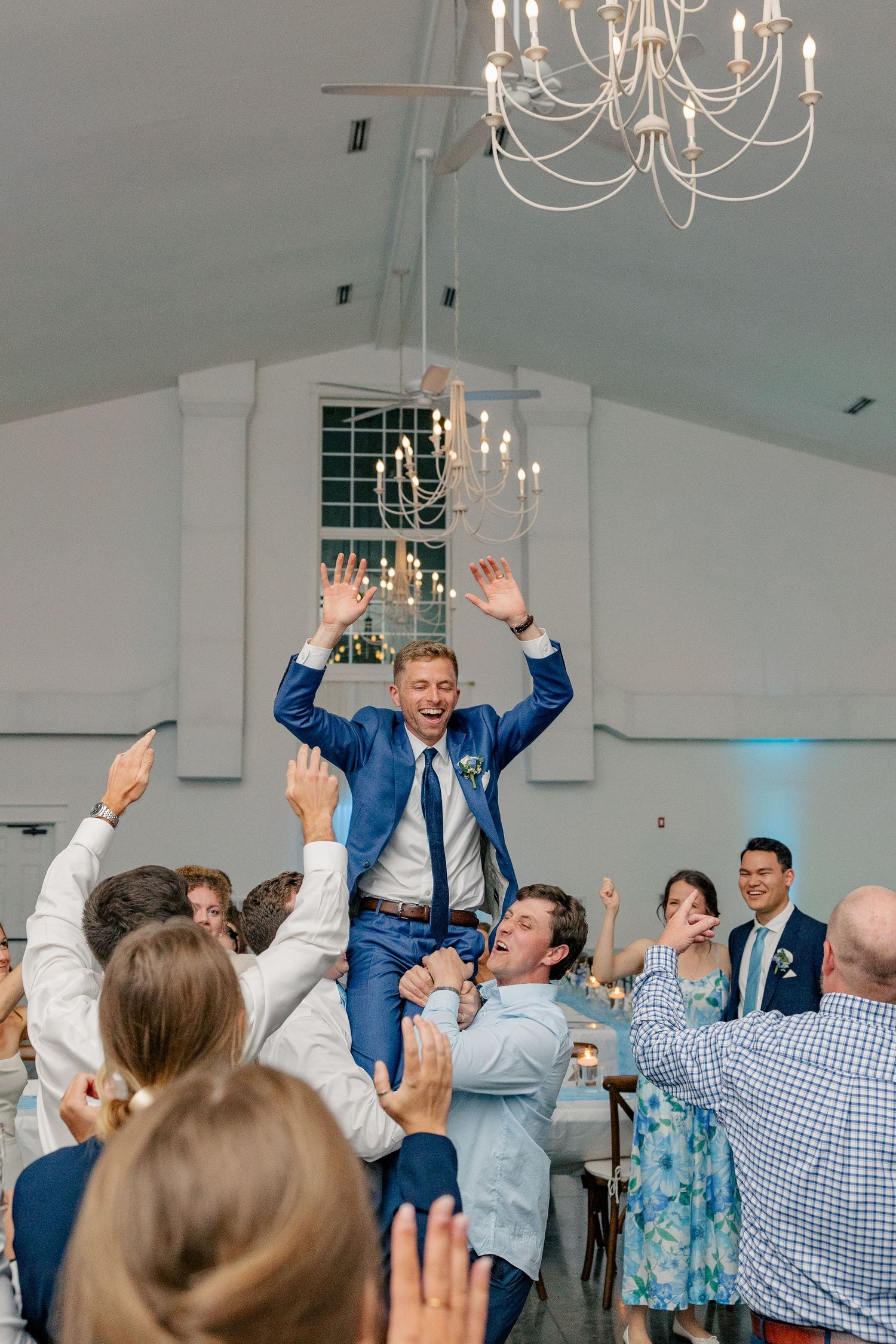 Groom in blue suit is lifted by groomsmen with hands raised in celebration at indoor wedding reception.