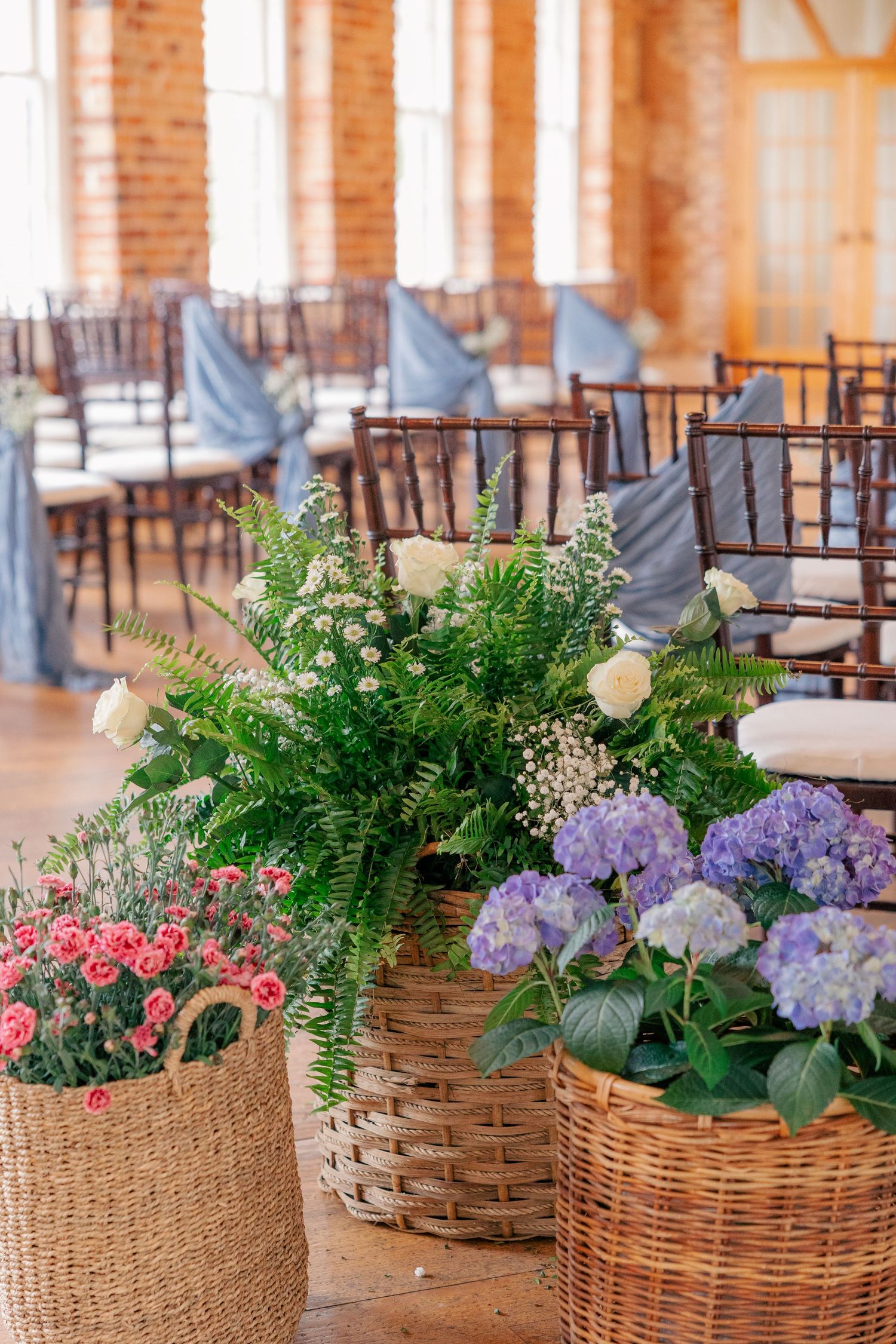 Wedding ceremony venue decorated with wicker baskets of colorful hydrangeas, ferns, and pink flowers beside chiavari chairs.