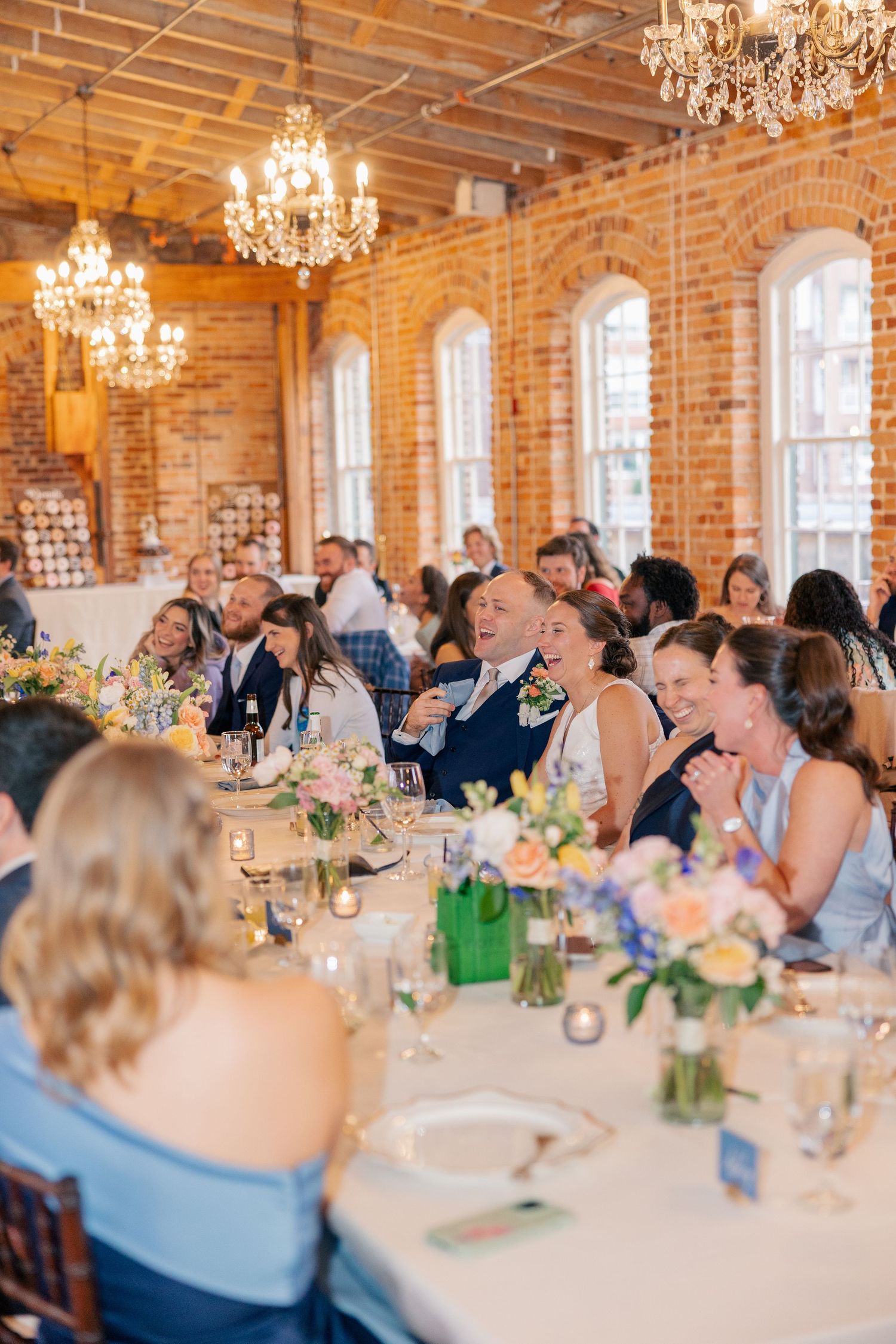 Wedding guests laugh and celebrate during a reception in a stunning brick venue with chandeliers.