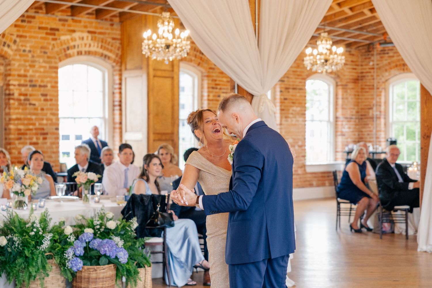 Groom in navy suit dancing with woman in gold dress in elegant brick ballroom with chandeliers and wedding guests.