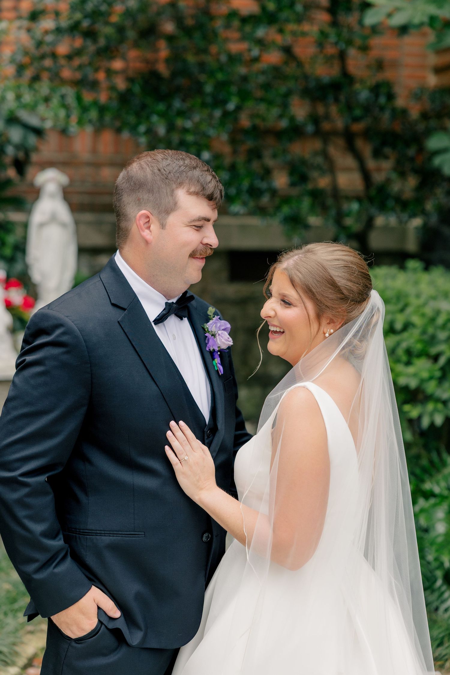 Bride and groom share a romantic outdoor moment, she smiles in a white veil dress as he gazes at her lovingly.