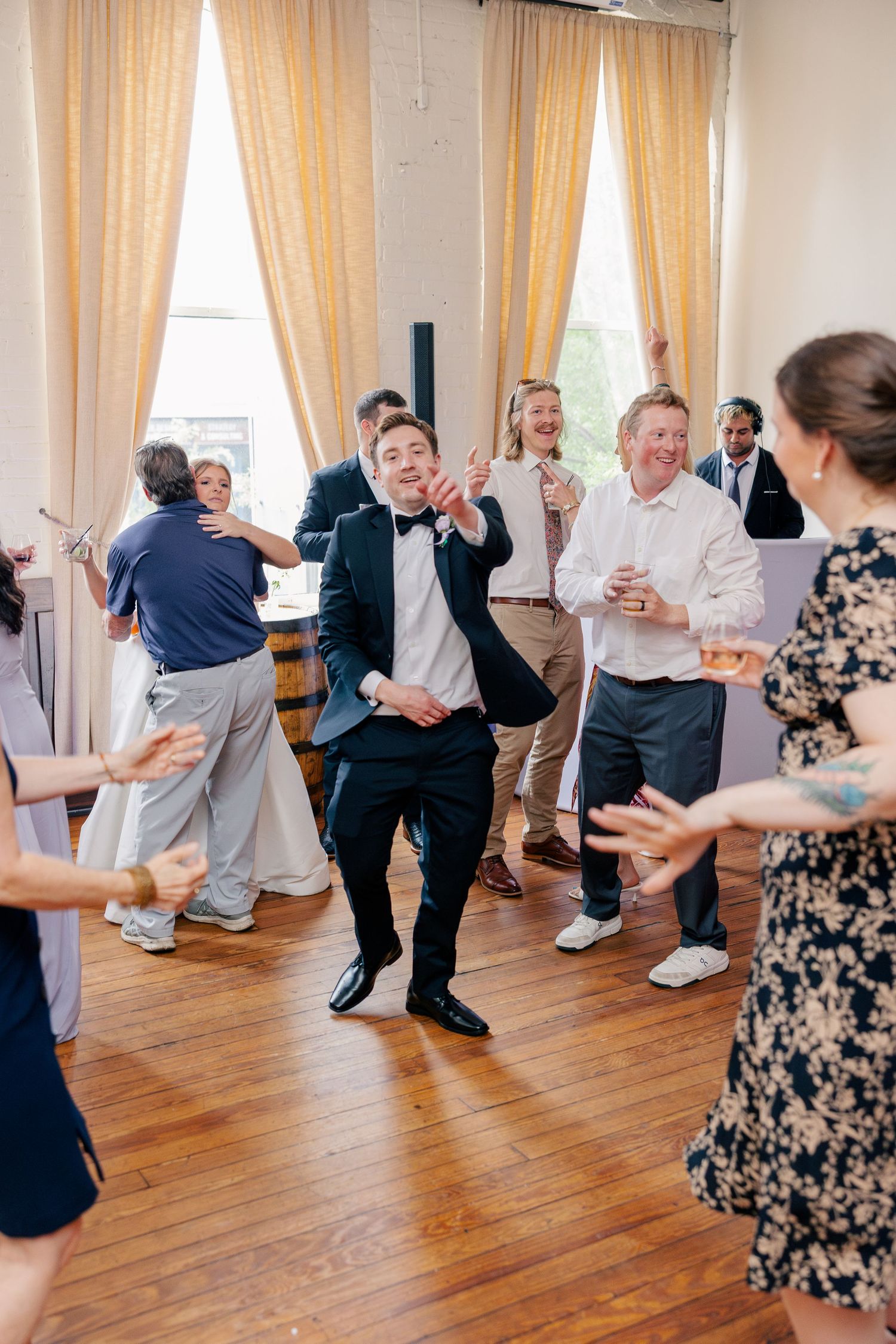 Groom in tuxedo dancing energetically with wedding guests on wooden dance floor.