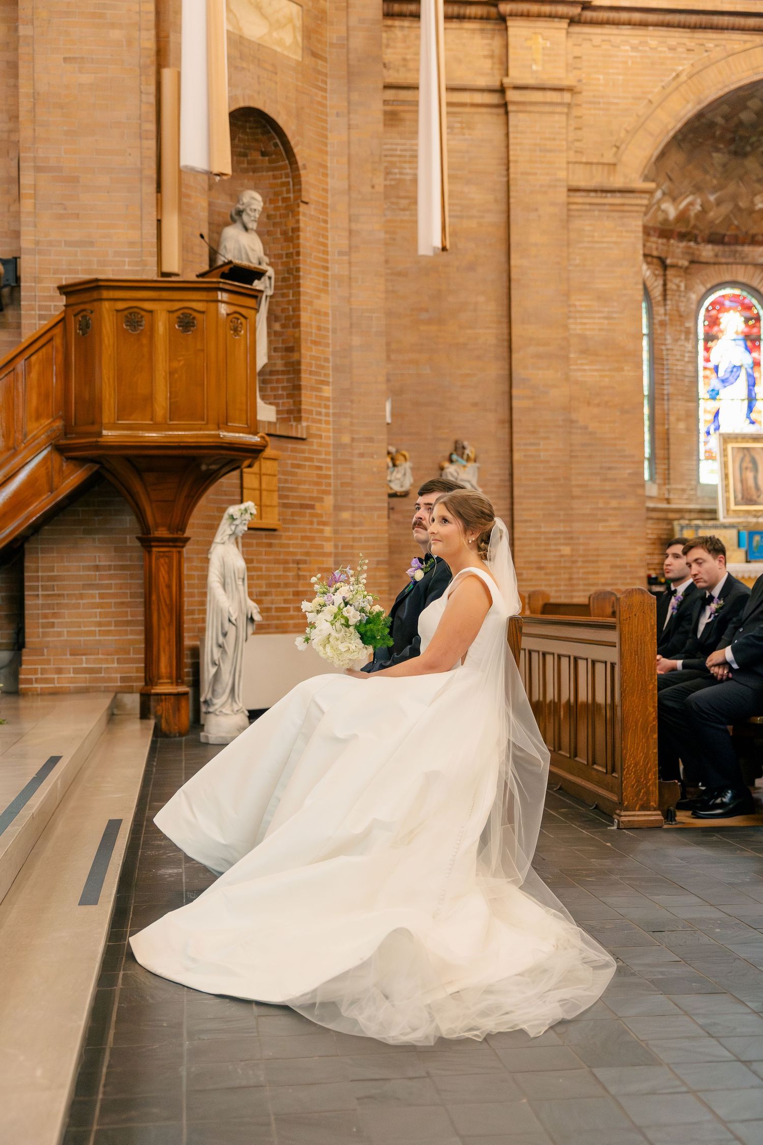 Bride in a flowing white gown and veil seated with groom during a Catholic church wedding ceremony.