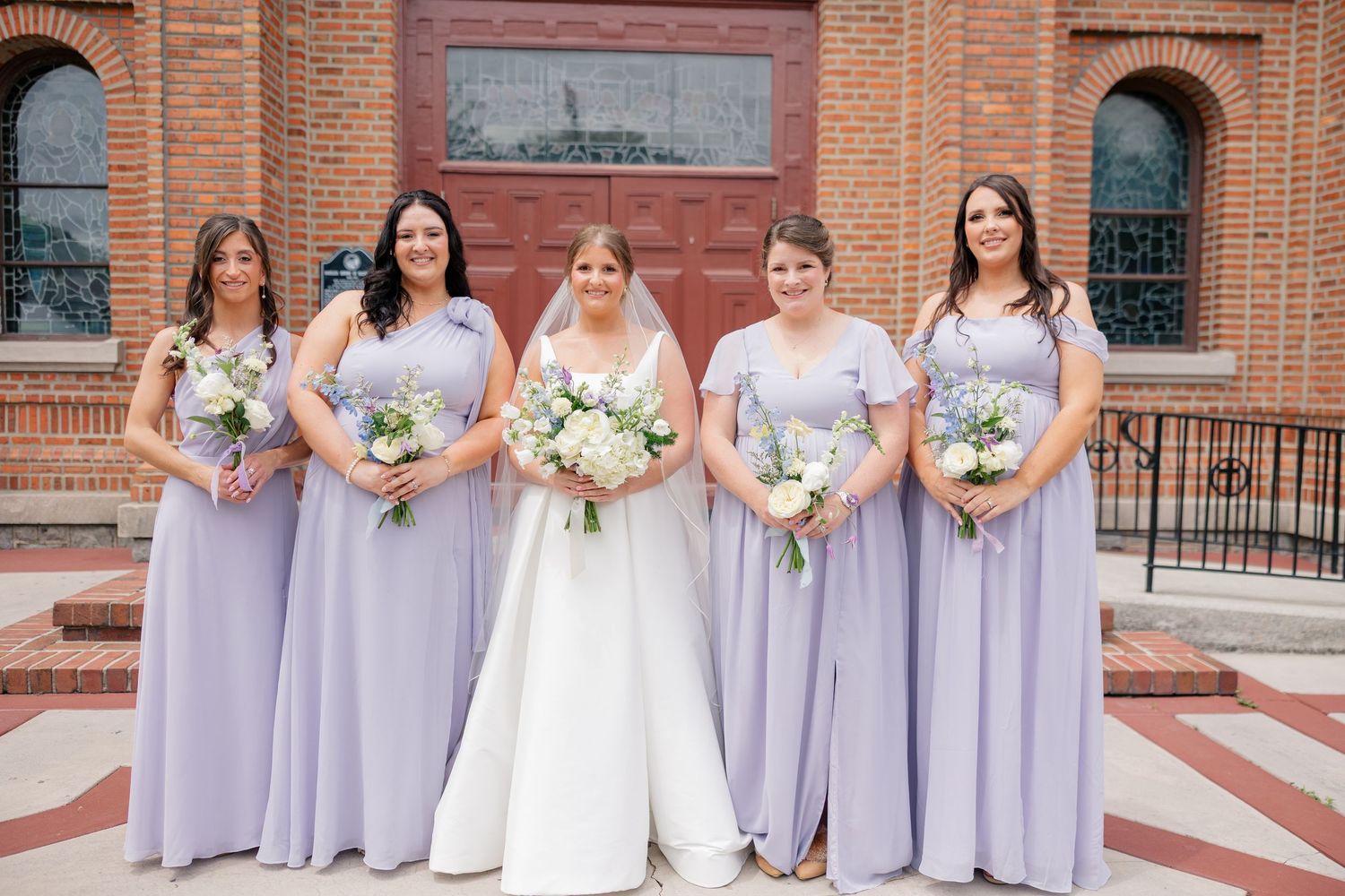 Bride in white gown poses with four bridesmaids in lavender dresses holding white bouquets before a brick church.