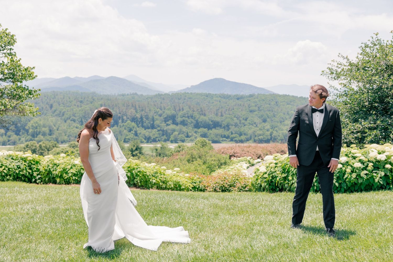 Bride and groom share a first look moment on a lush green lawn with scenic mountain views in the background.