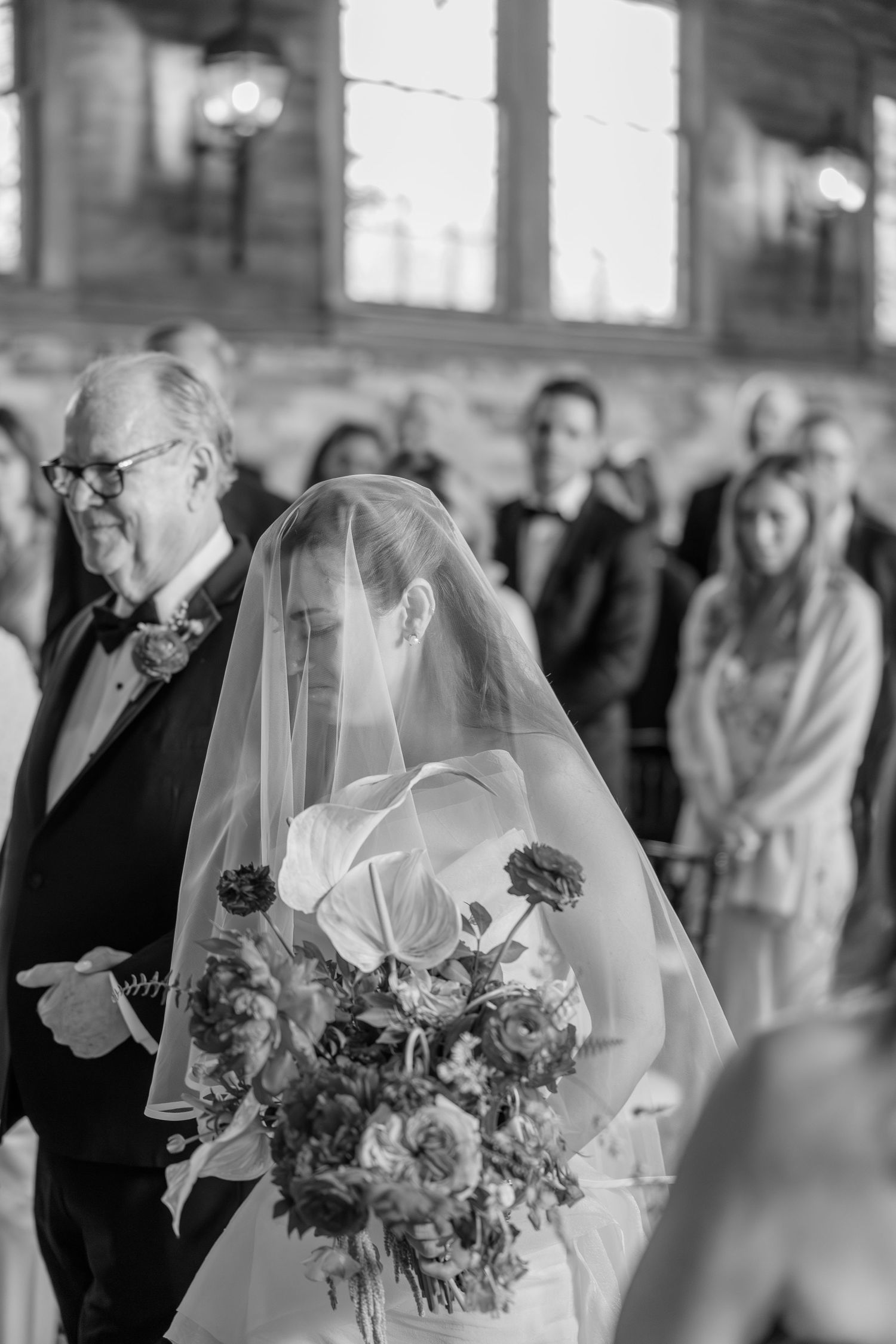 Black and white photo of bride walking down the aisle with her father holding a lush floral bouquet.