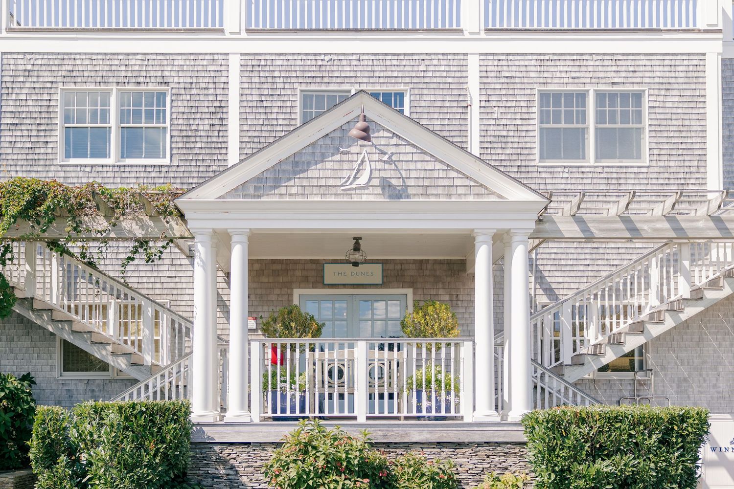 Charming coastal shingle-style building with white portico entrance, twin staircases, and lush greenery.