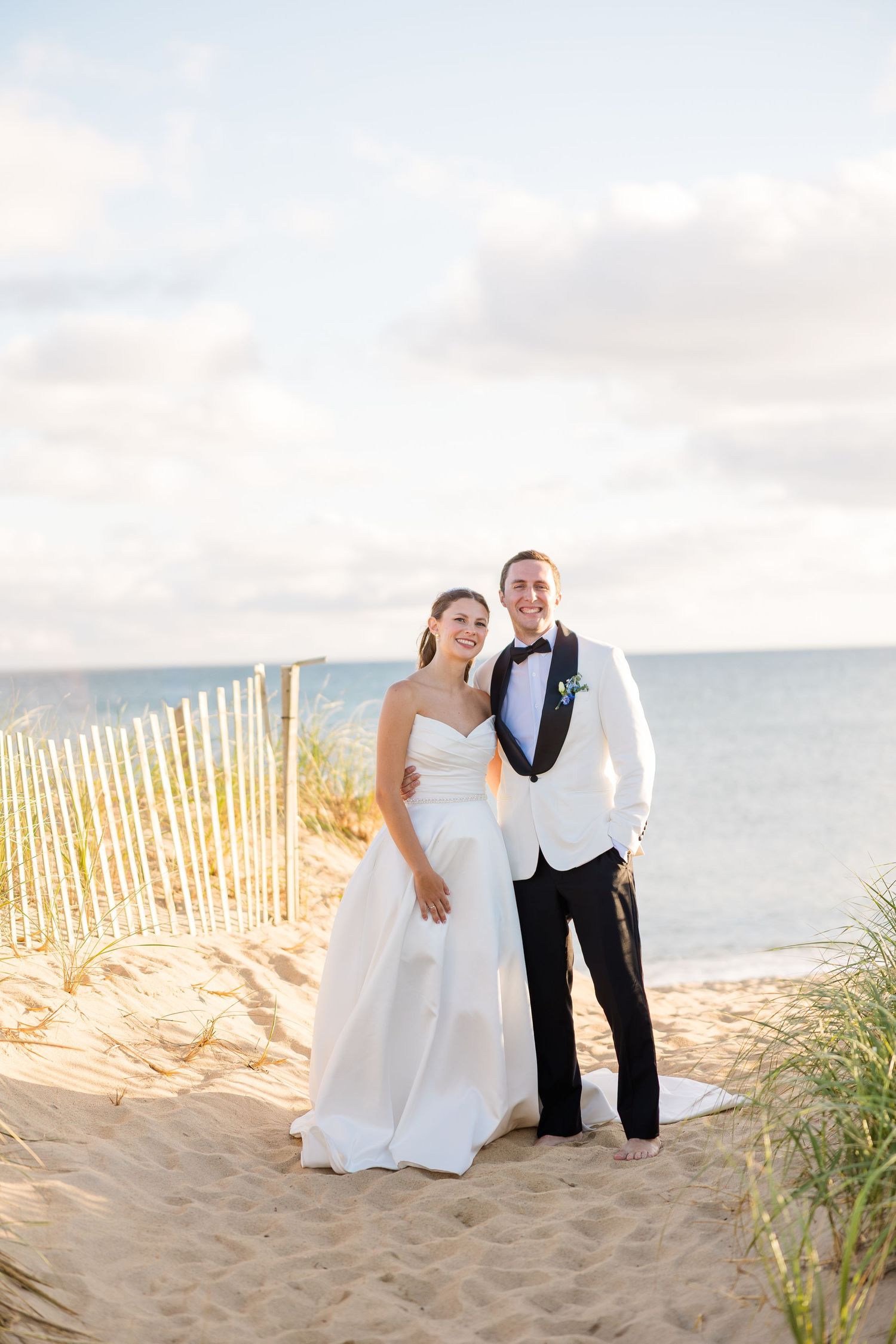 Bride and groom pose together on sandy beach path with ocean and wooden fence in golden hour light.