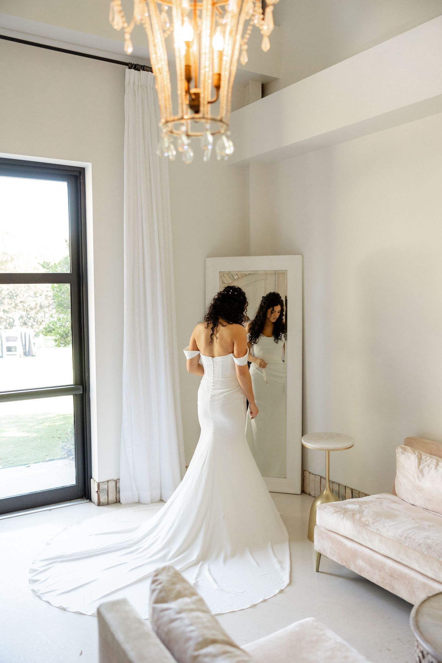 A bride in a stunning white mermaid wedding gown with long train looks at herself in a mirror in an elegant room.