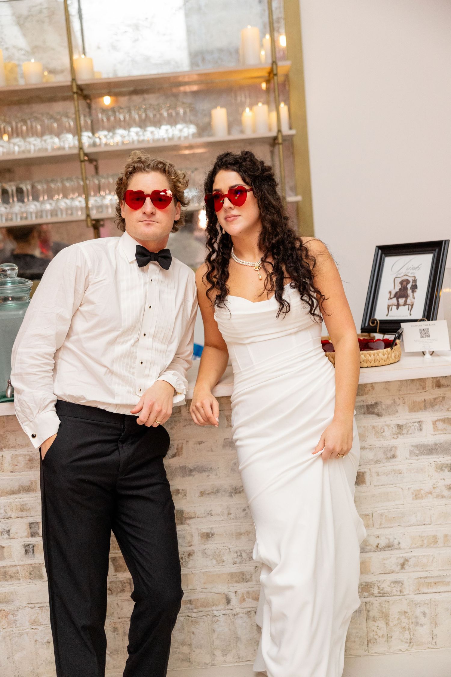 Newlyweds wearing red heart sunglasses pose playfully against exposed brick wall with glassware shelving.