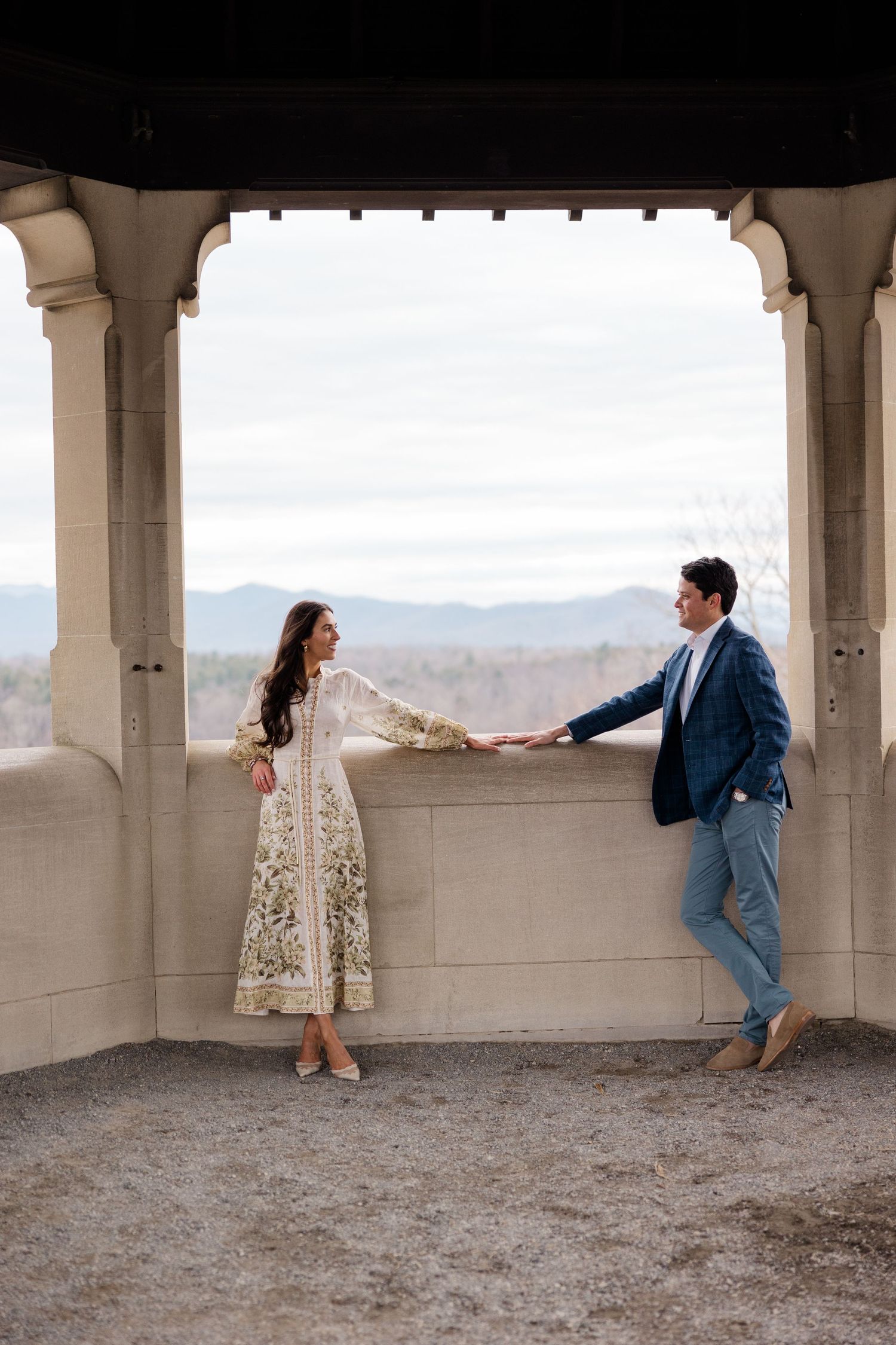 A couple in formal attire shares a romantic moment on an outdoor stone terrace with mountain views in the background.