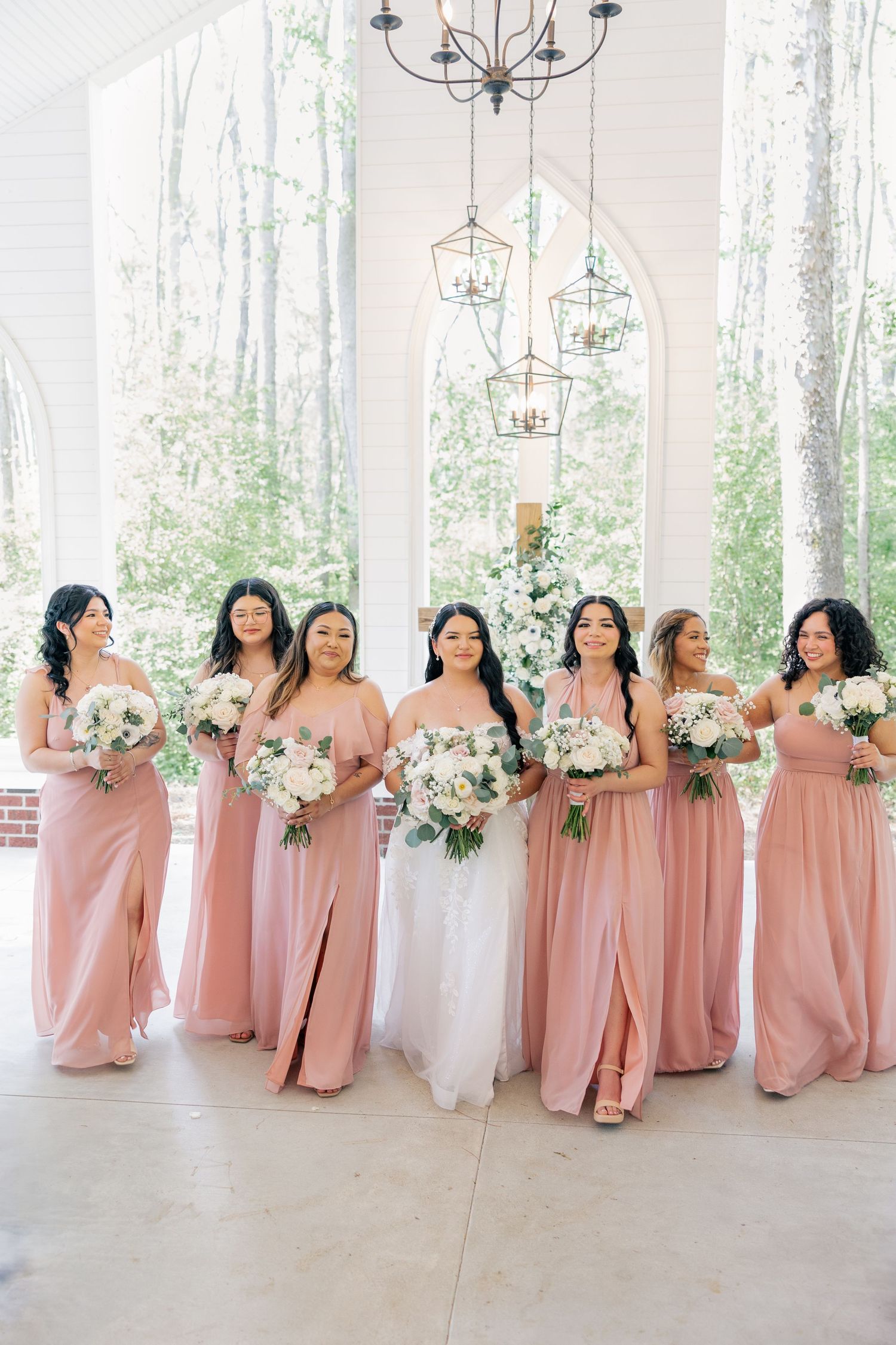 Bridesmaids in blush pink dresses hold white floral bouquets while posing in a bright venue.