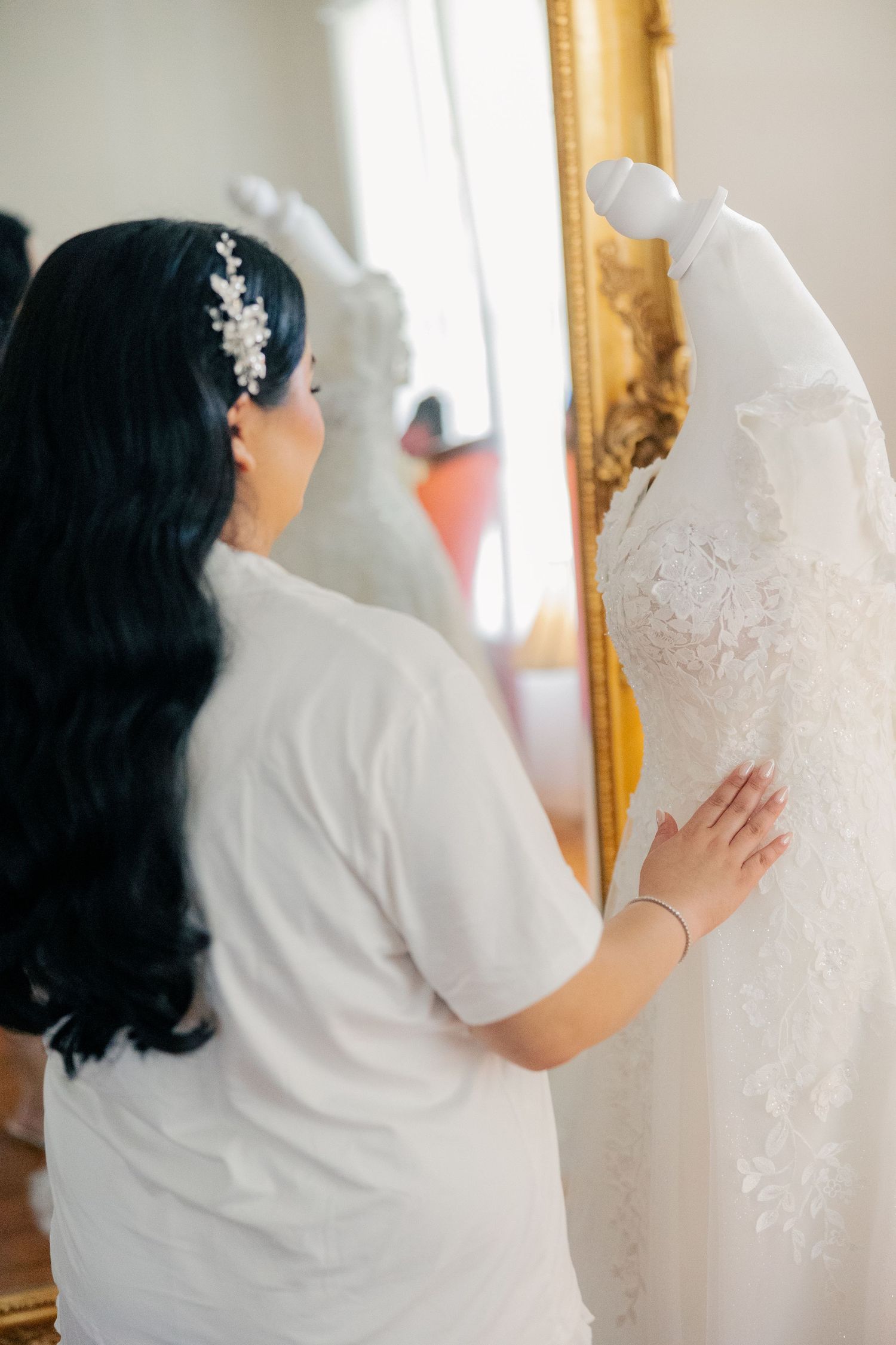 A wedding dress being inspected on a hanger against an ornate gold-framed mirror.