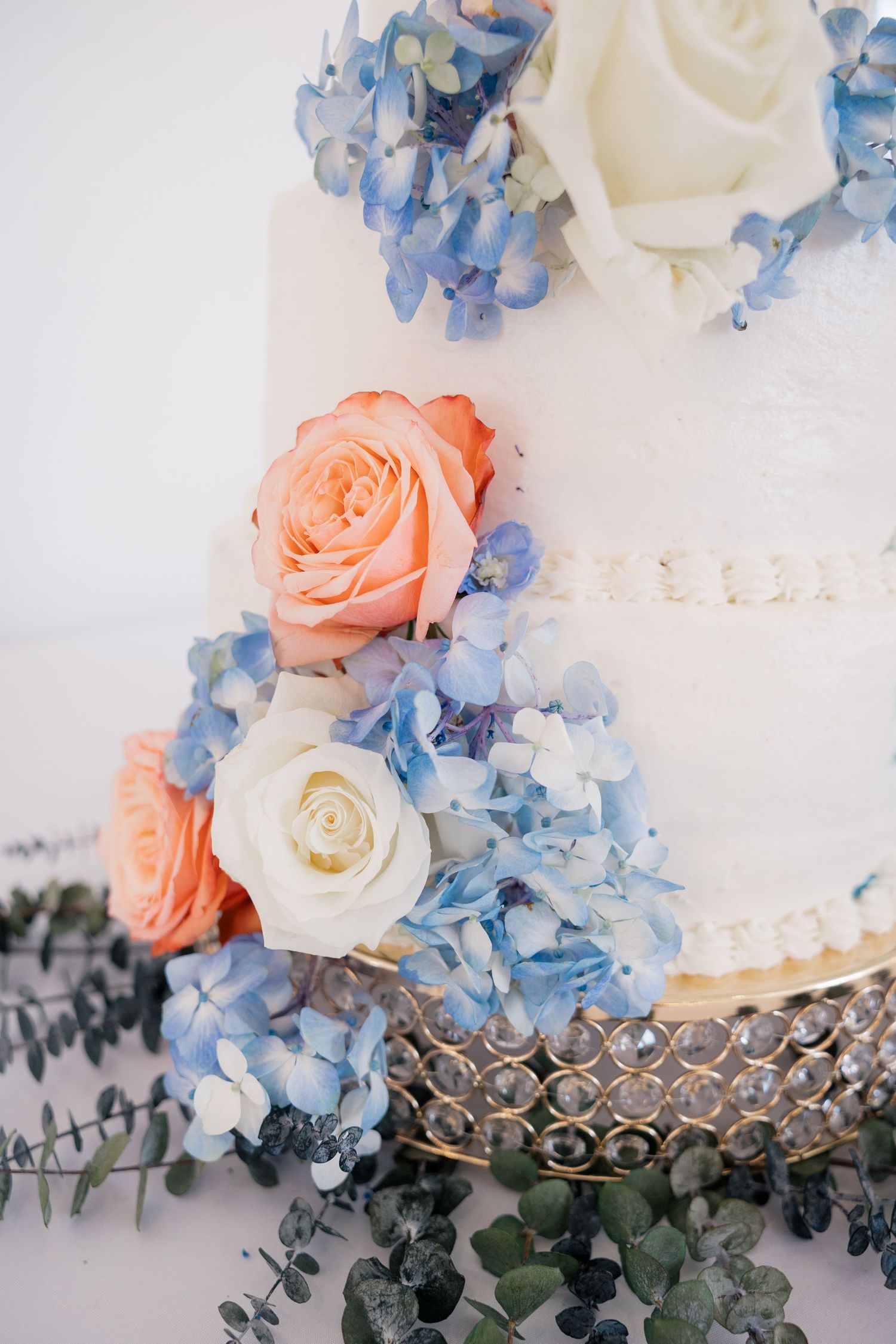 Wedding cake decorated with peach roses, white flowers and blue hydrangeas on a silver stand.