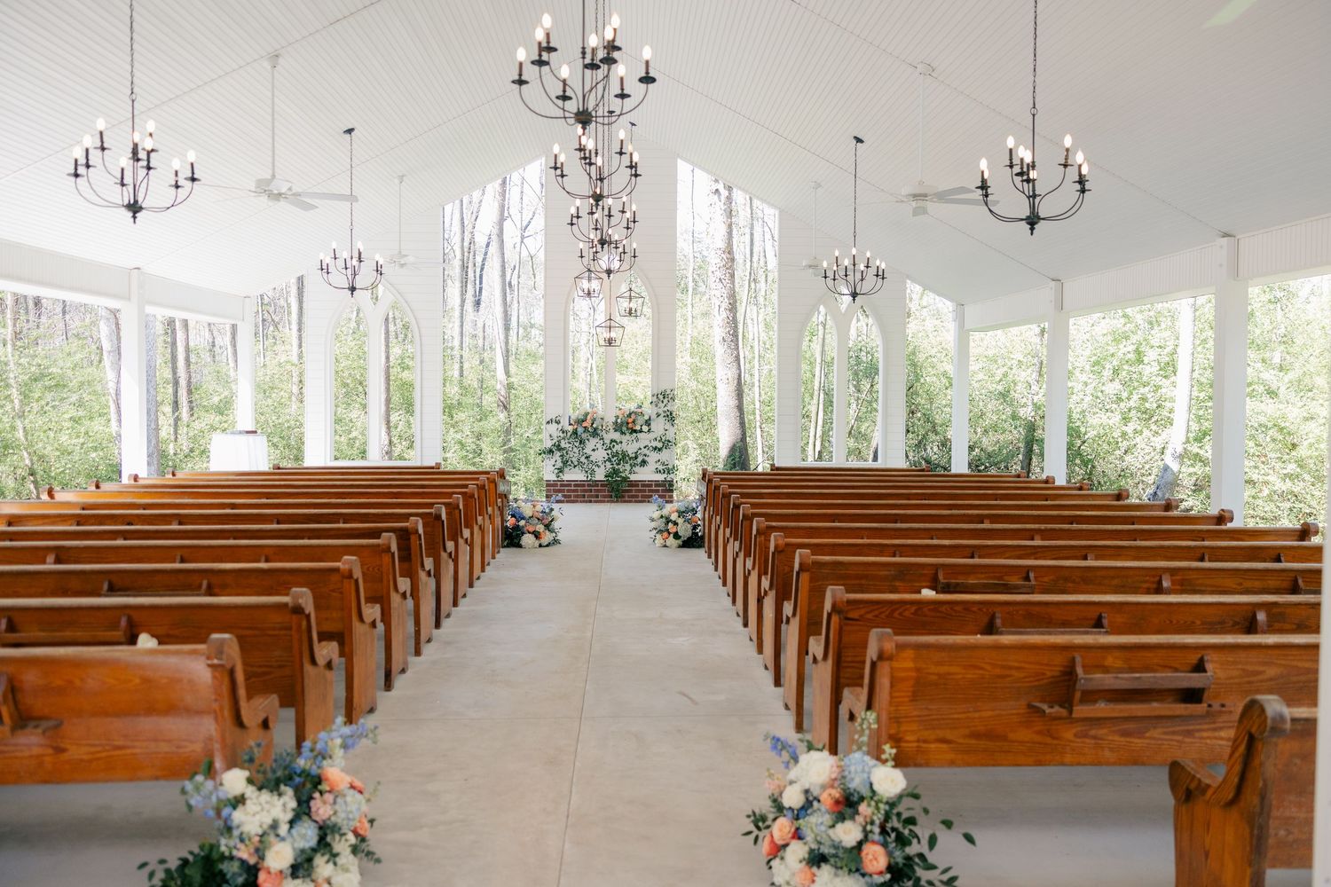 Modern white chapel interior with chandeliers and large windows overlooking woods.