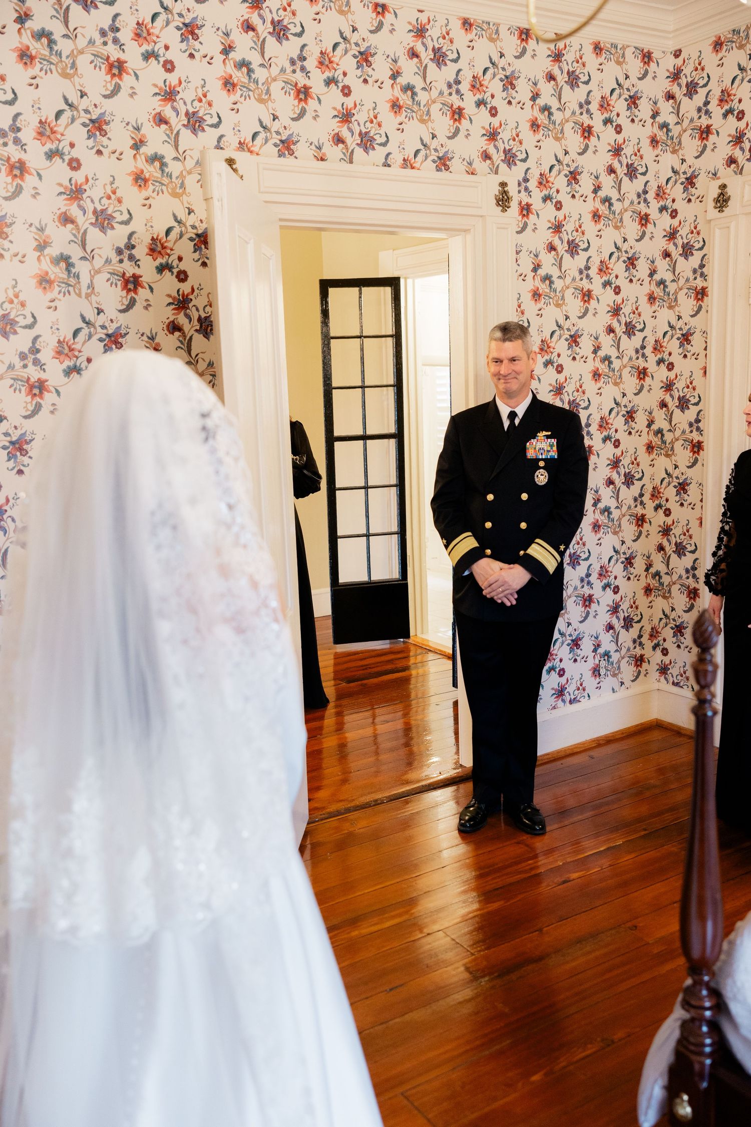 A wedding dress and uniformed figure stand in a floral wallpapered room with hardwood floors.