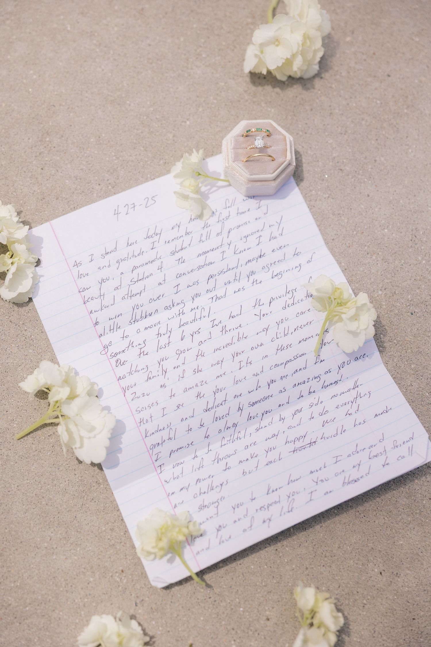 A handwritten letter on white paper surrounded by scattered white flowers and petals on a gray surface.