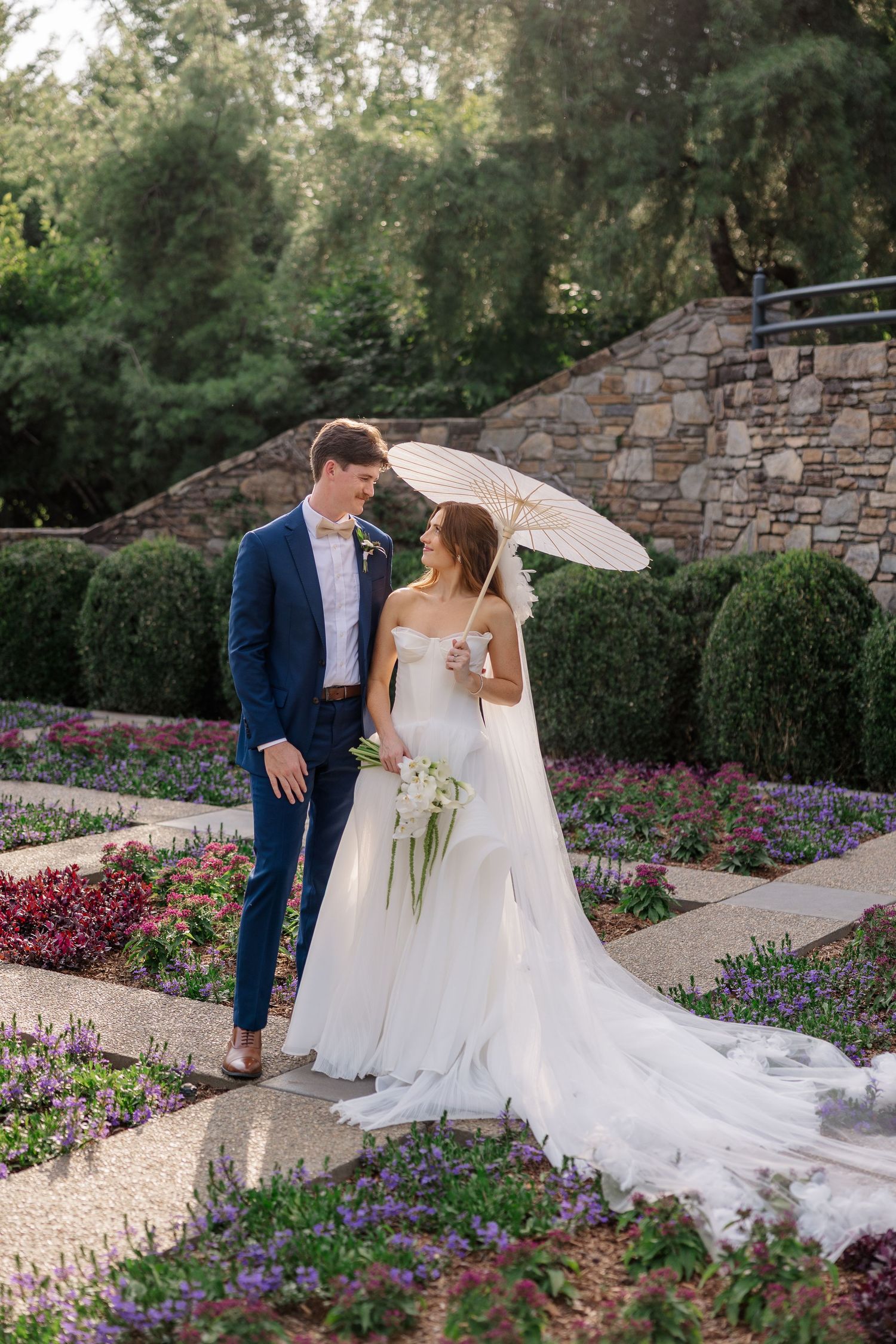 A wedding couple poses in a beautiful garden with a white parasol and stone wall backdrop.