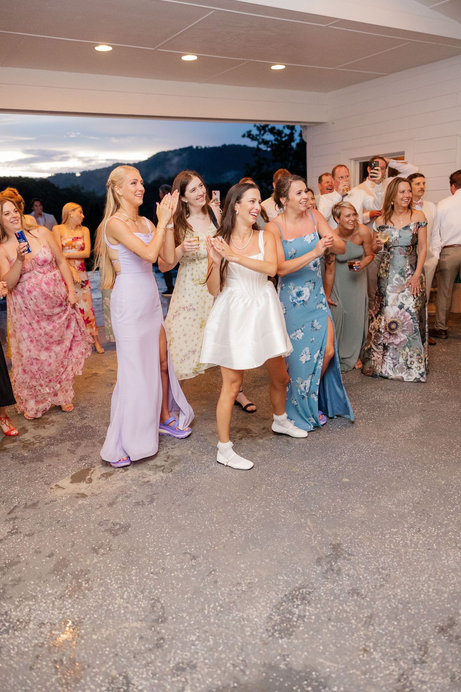 Wedding guests in colorful dresses dance in a line on the dance floor at a reception.