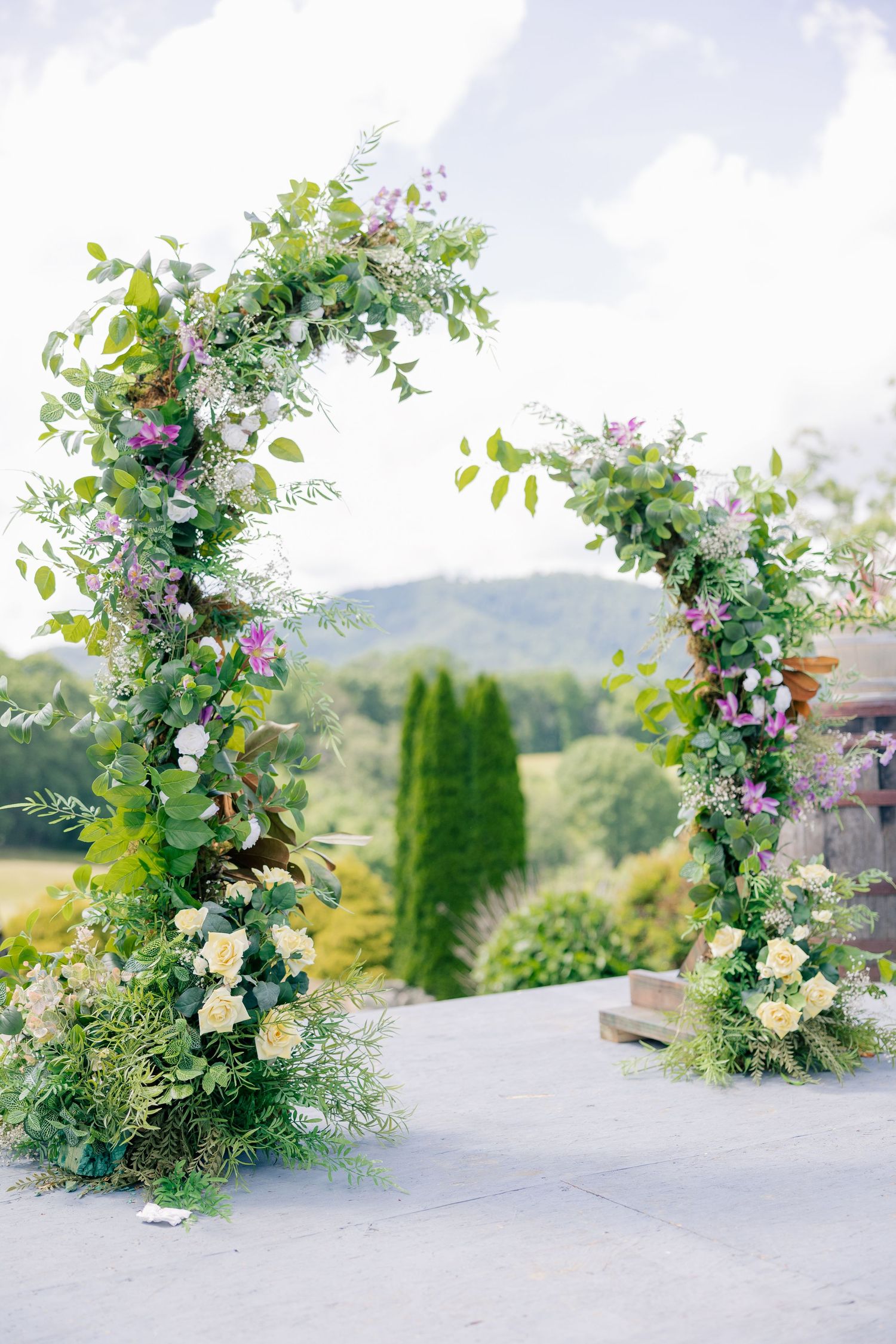 A floral arch with purple and white blooms frames a mountain view on a sunny day.