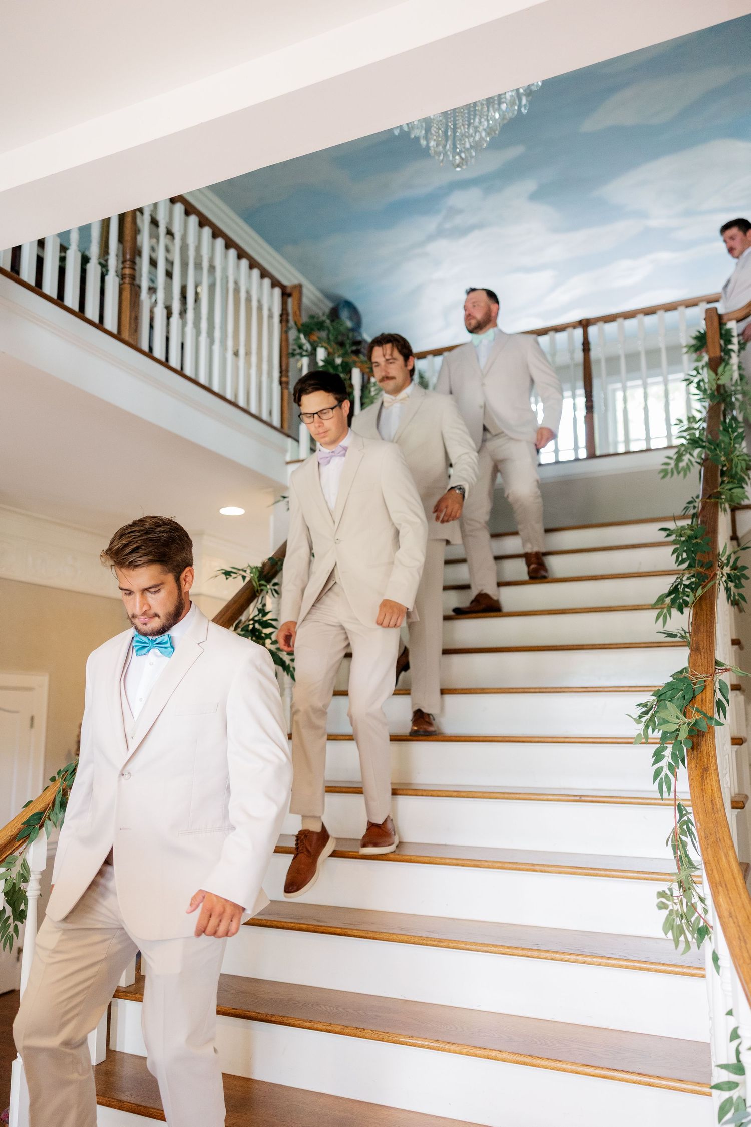 Groomsmen in light beige suits descending an elegant staircase decorated with greenery.