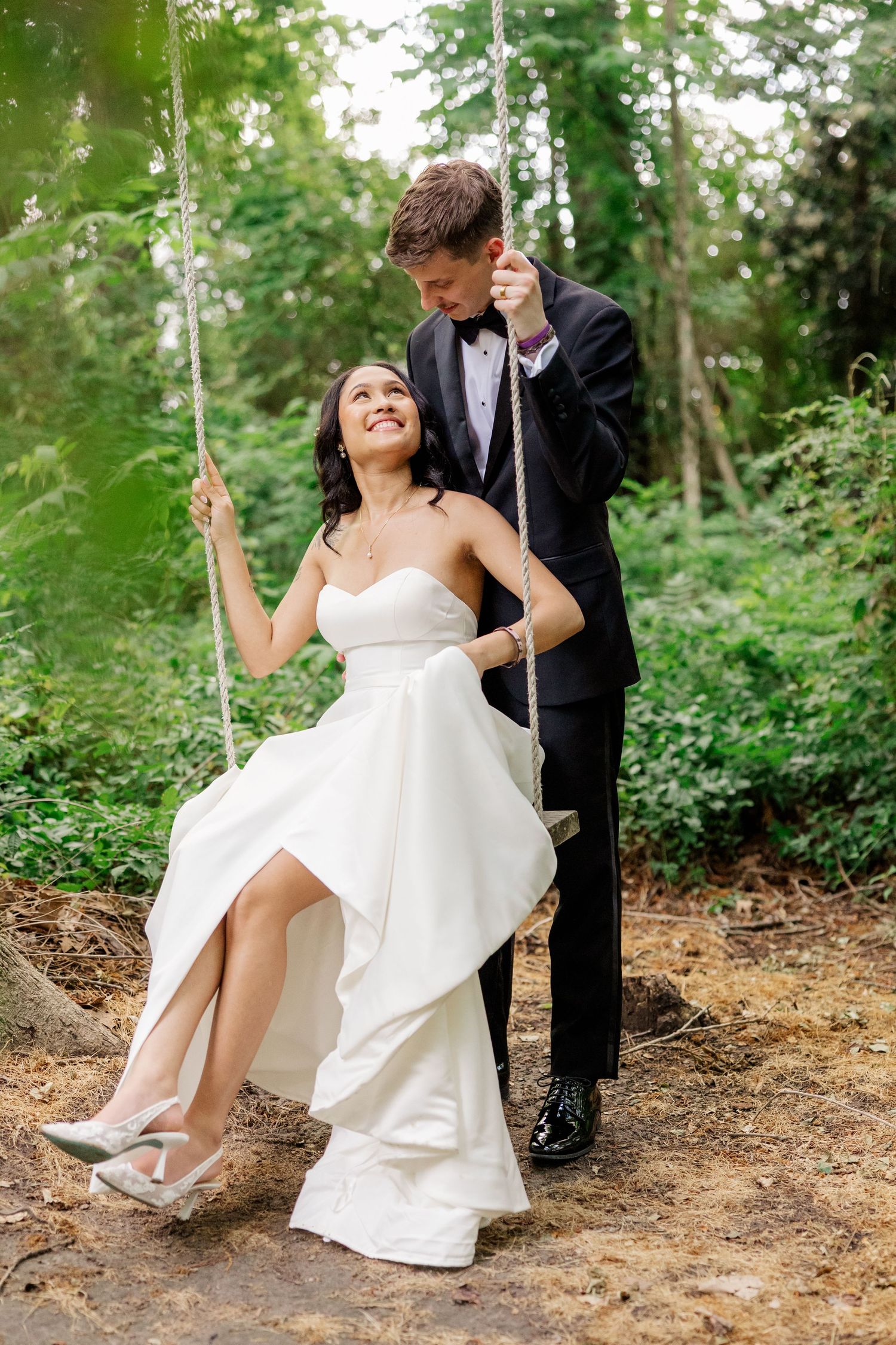 Wedding couple playfully poses on a rustic wooden swing in a forest setting.