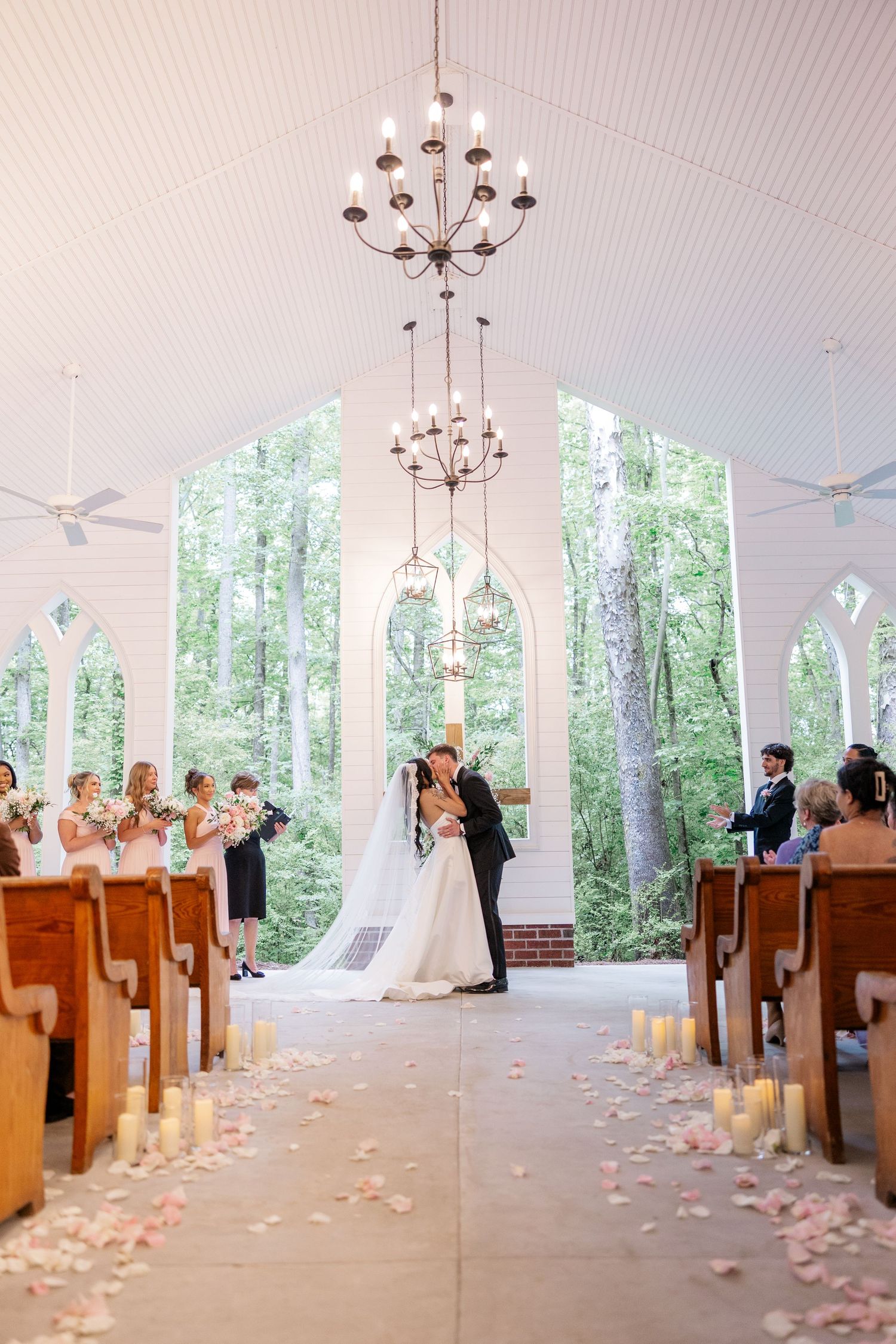 A white chapel interior with wooden pews and chandeliers decorated for wedding ceremony with rose petals.