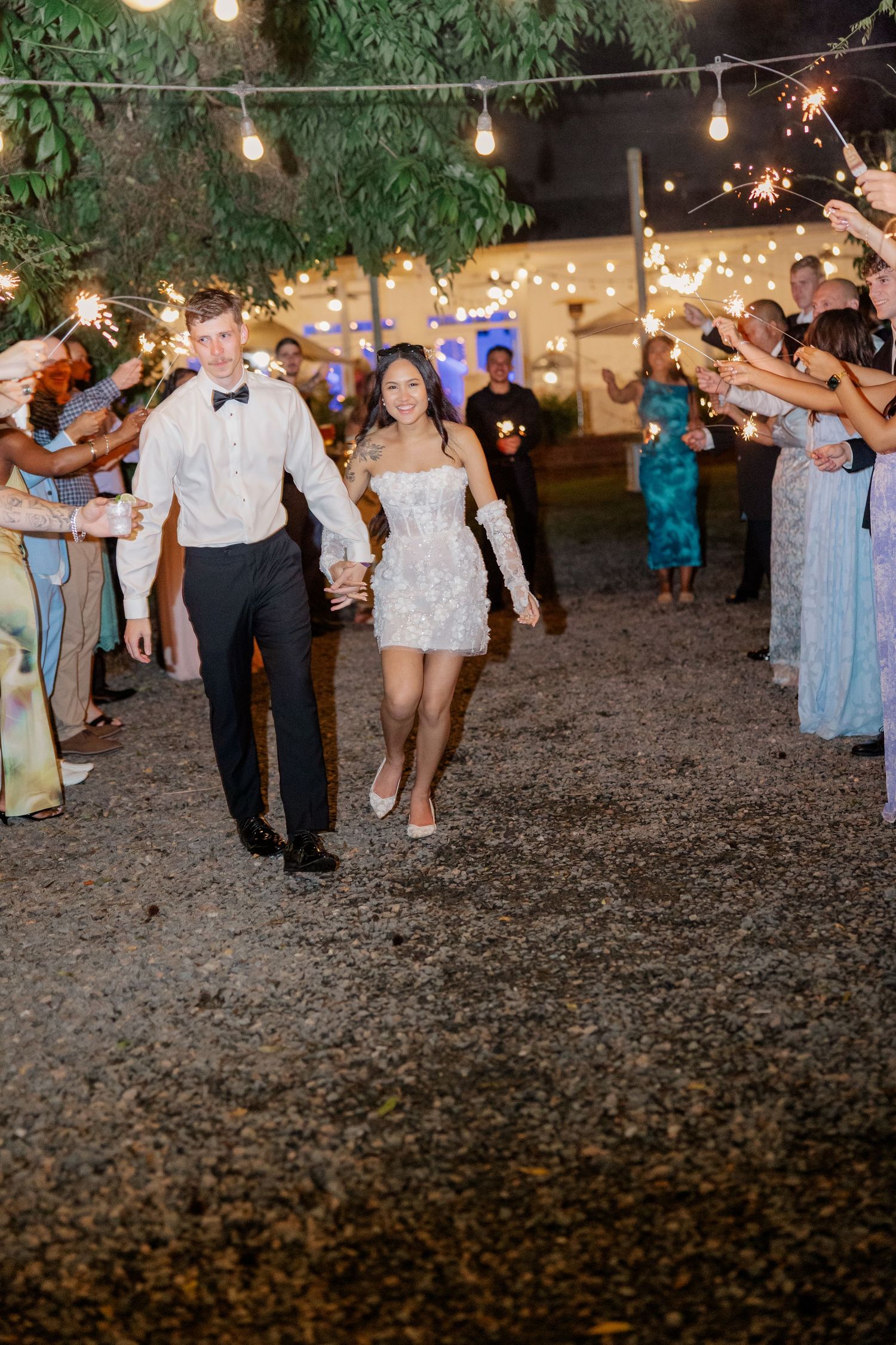 Wedding party and guests dancing outdoors at night under strings of glowing bistro lights.