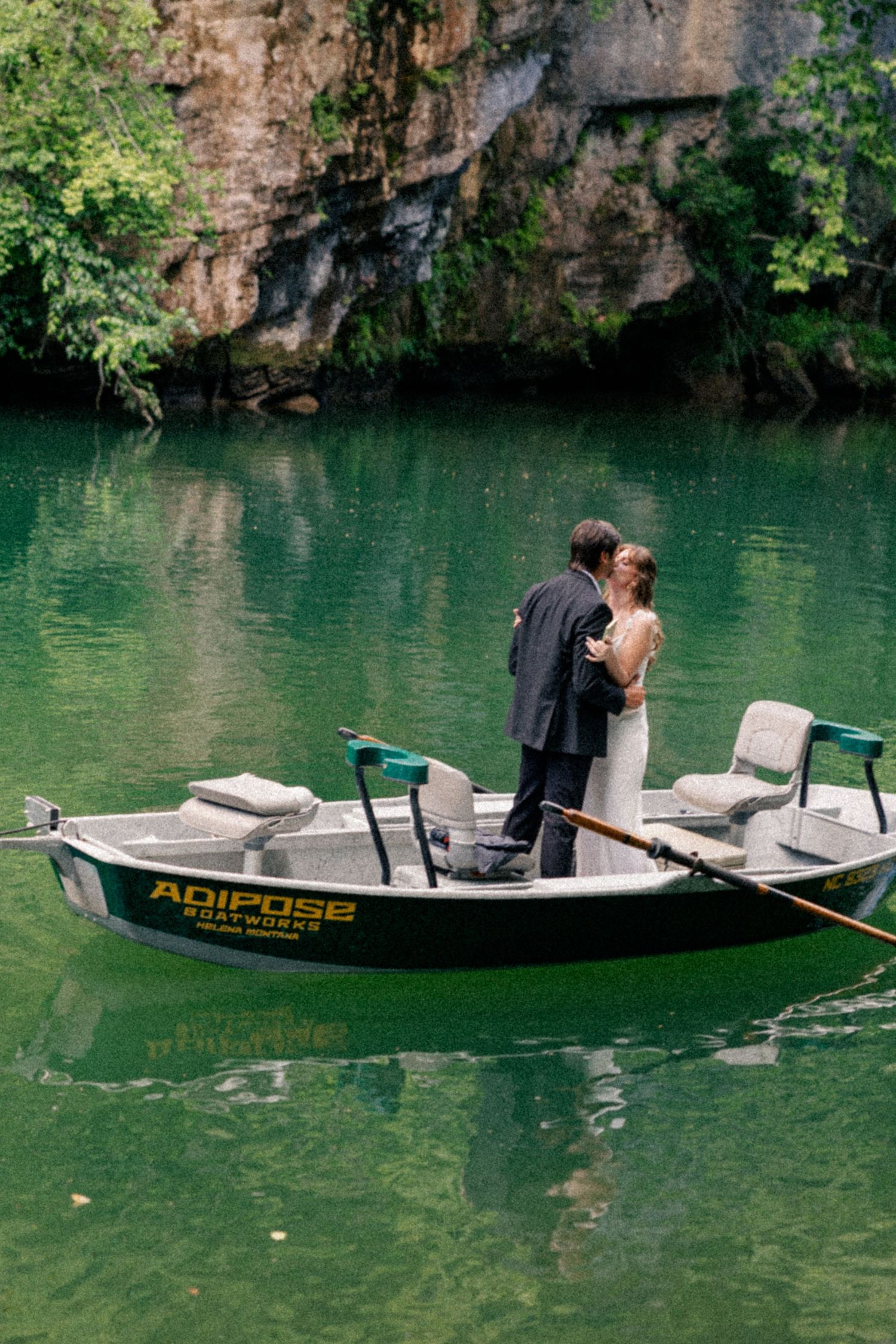 Bride and groom share a kiss standing in a boat on a stunning emerald green river.
