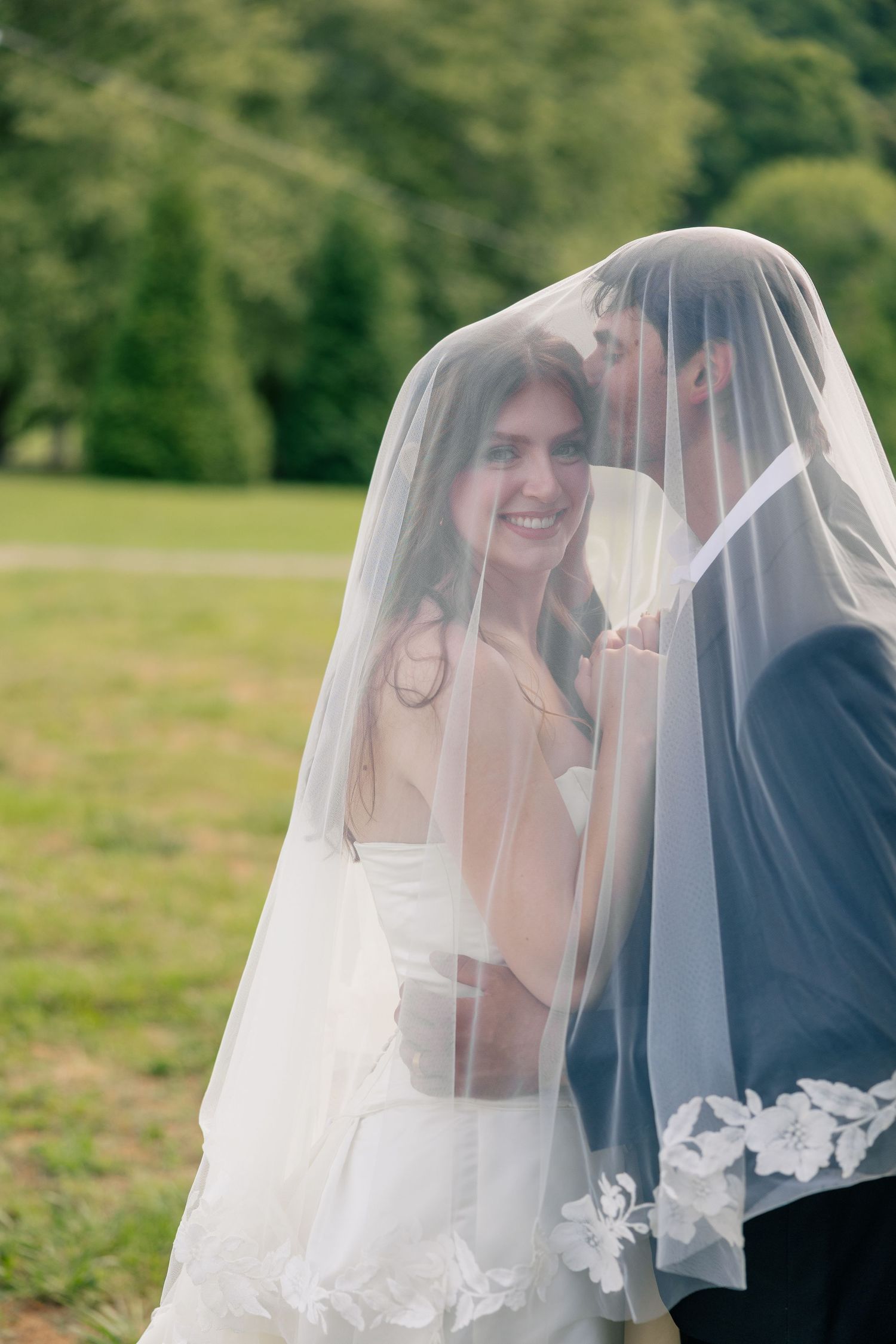 Smiling bride and groom share an intimate moment hidden beneath her lace-trimmed cathedral wedding veil.