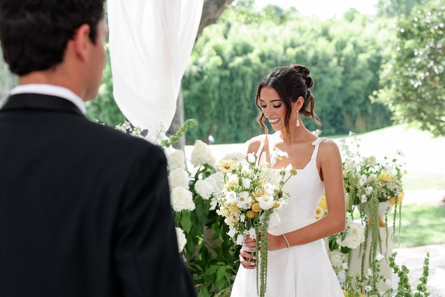 Bride in white gown holding yellow and white bouquet smiles at groom during outdoor wedding ceremony.