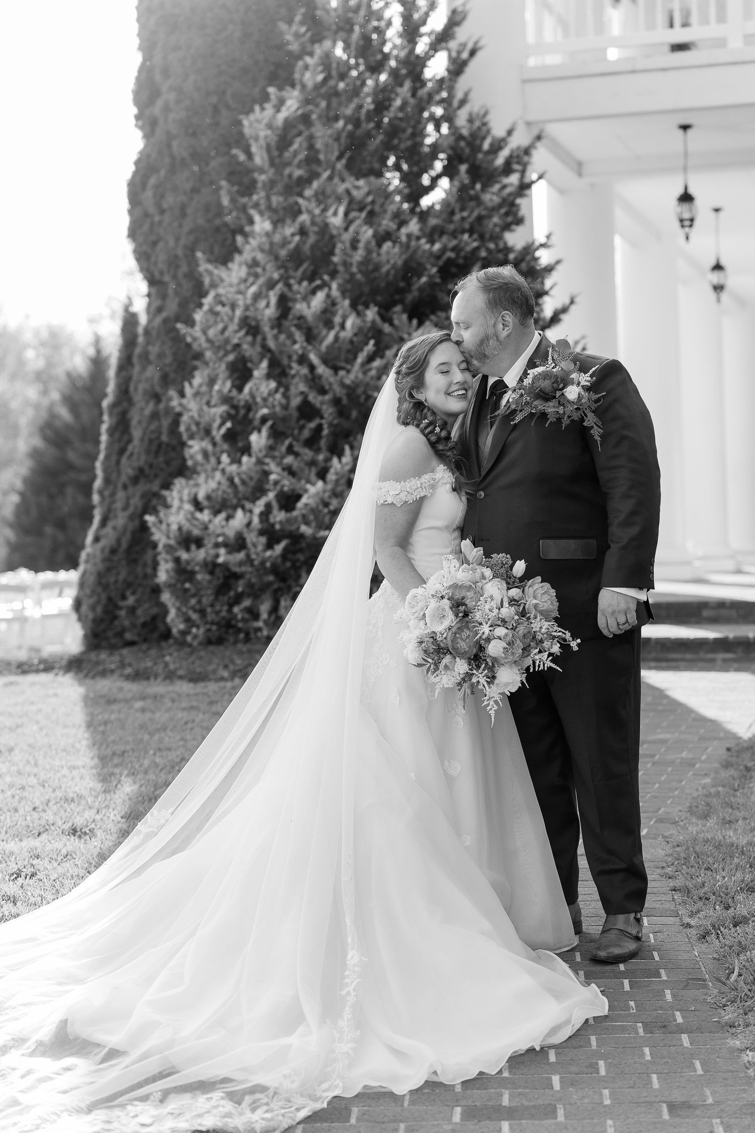 Black and white photo of a bride and groom sharing a tender moment outdoors, with her flowing veil and bouquet on display.