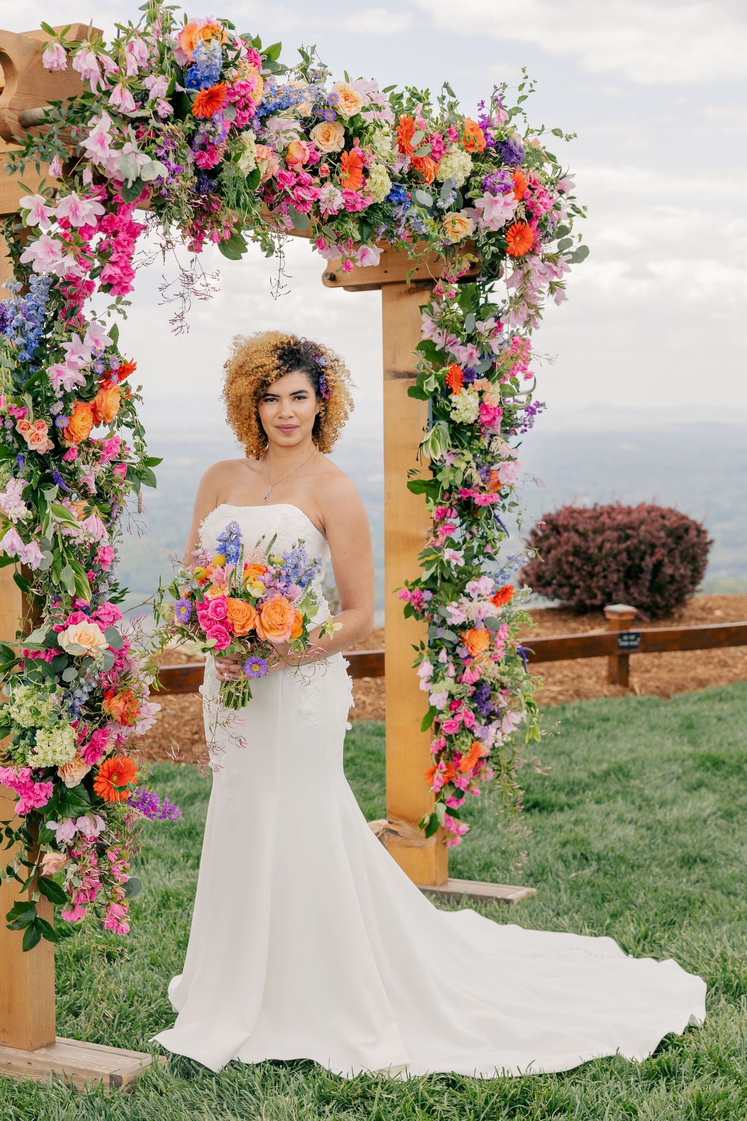 Bride in white gown holding colorful bouquet under vibrant floral arch with mountain views behind her.
