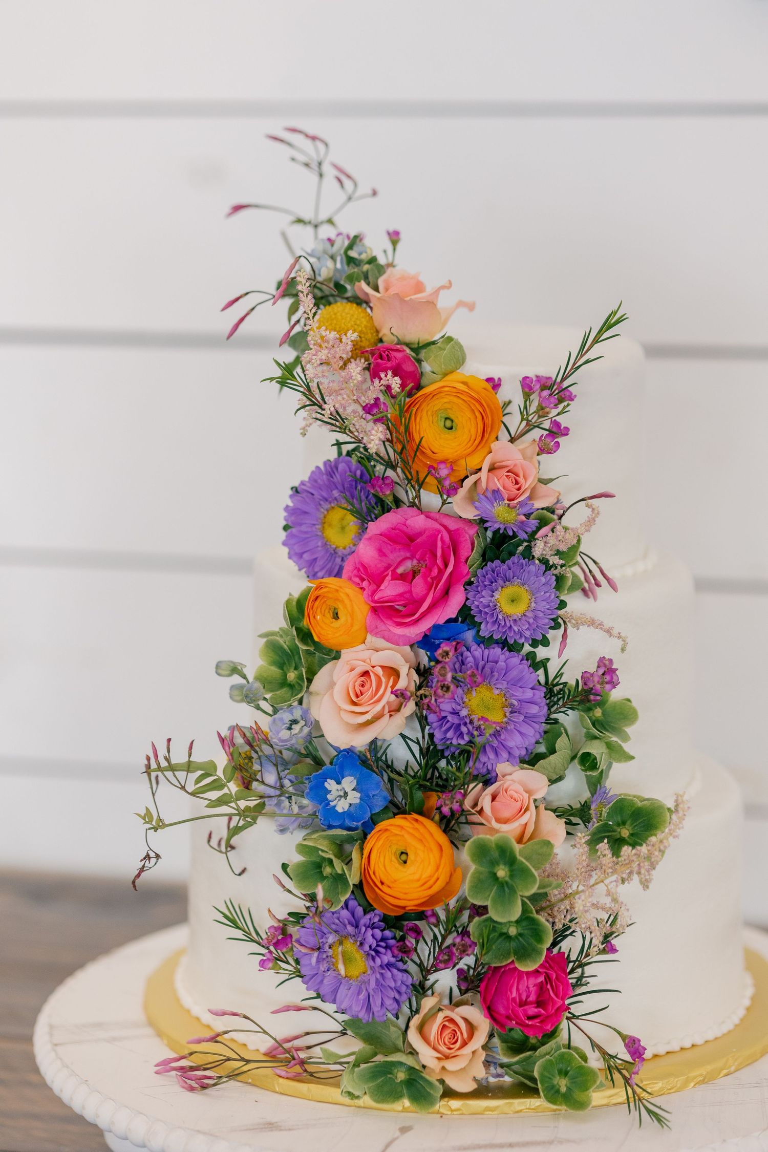 Elegant white wedding cake adorned with colorful wildflowers including pink roses, orange ranunculus, and purple asters.