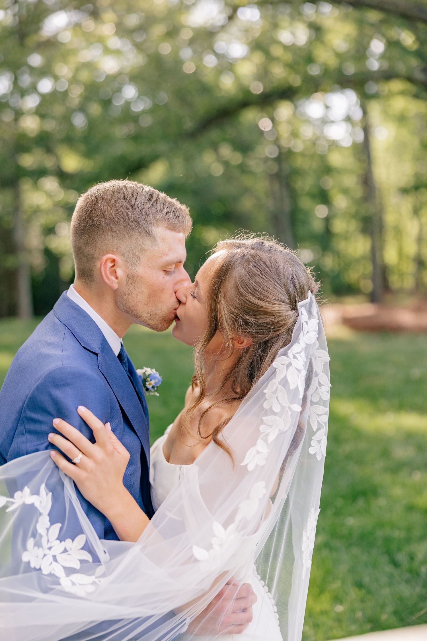 Groom in blue suit kisses bride in lace veil as she embraces him in lush green outdoor setting.