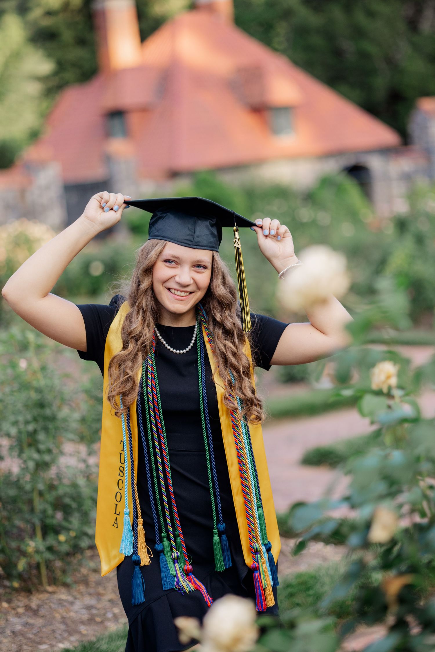 A graduate in cap and gown with colorful honor cords poses playfully outdoors during a graduation photoshoot.