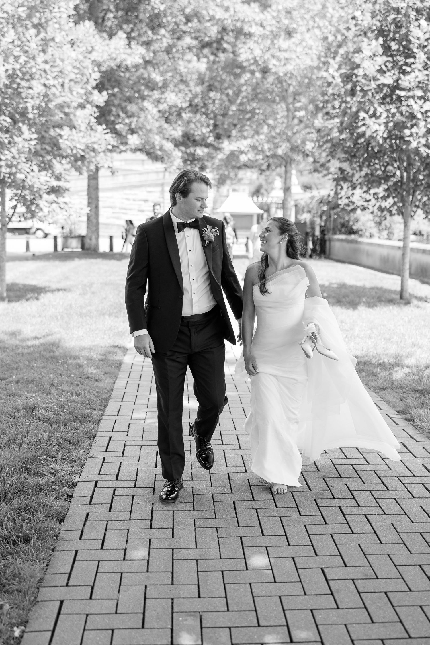 Bride and groom walking hand in hand on brick path surrounded by trees in black and white photo.