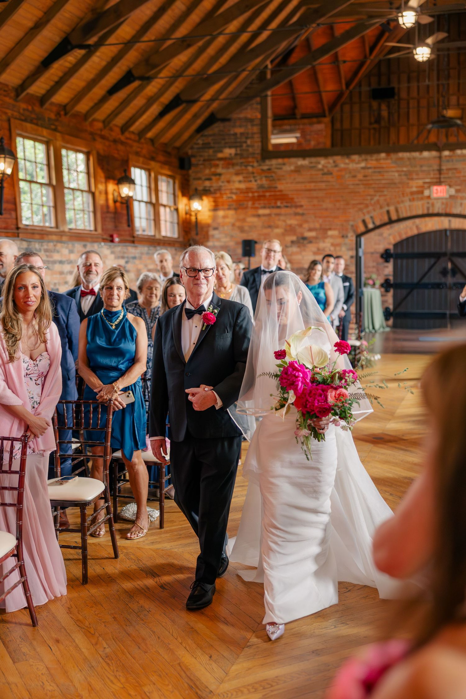Bride holding pink bouquet walks down aisle with escort in rustic brick barn wedding venue.