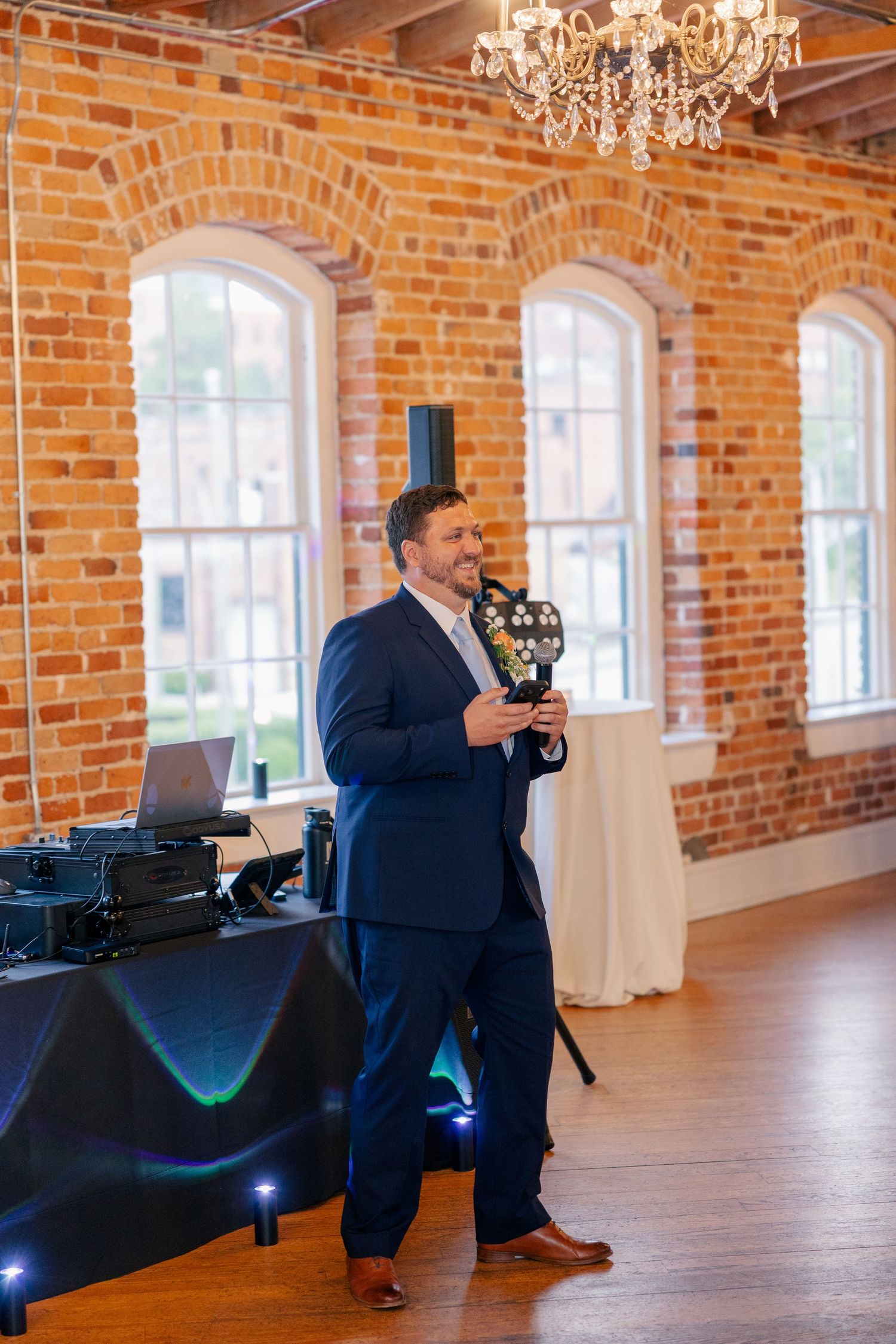 Wedding DJ speaks into a microphone in a rustic brick ballroom with crystal chandeliers overhead.