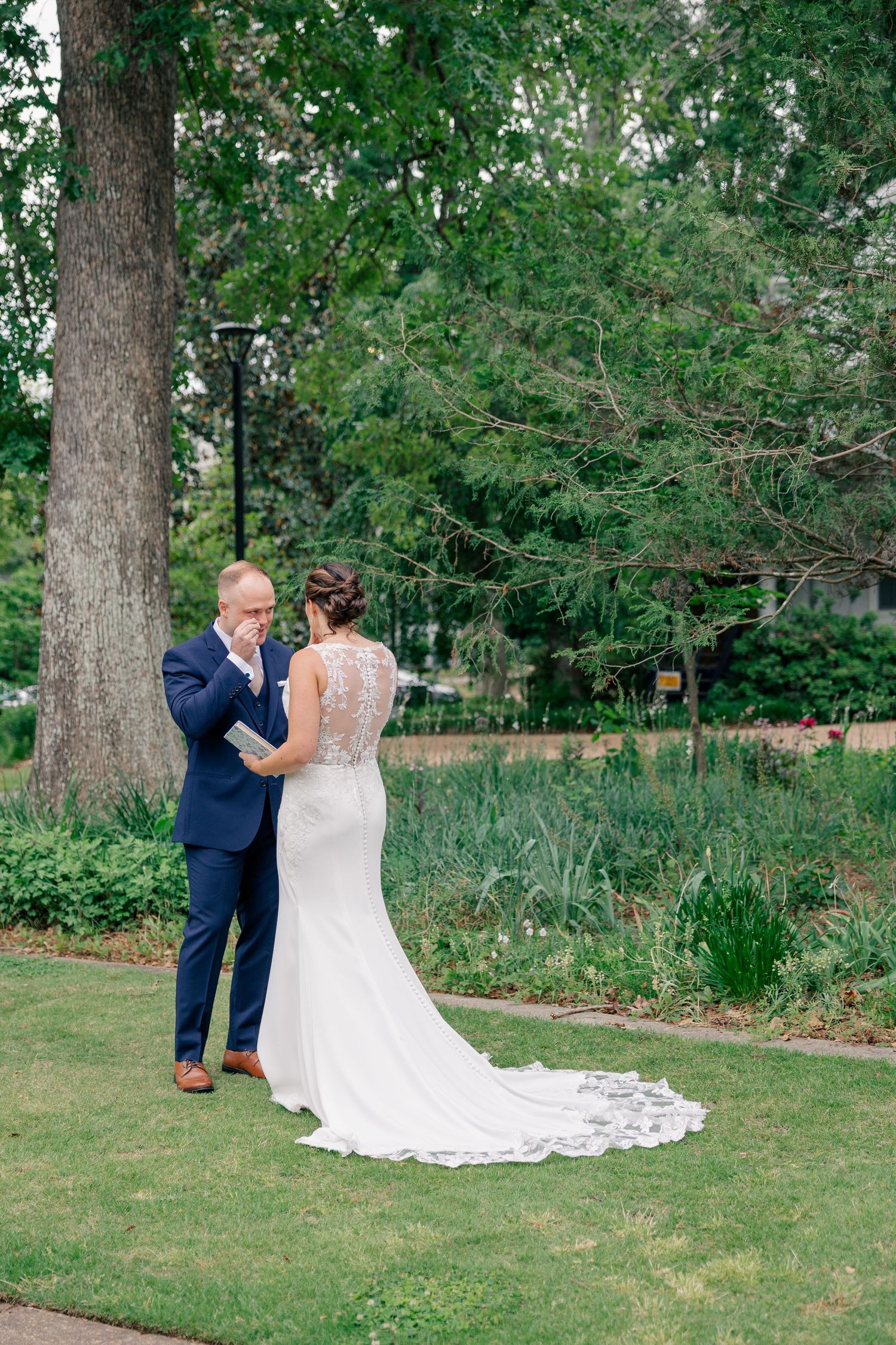 Emotional groom reads vows to bride in lace-back gown during outdoor first look in green park.